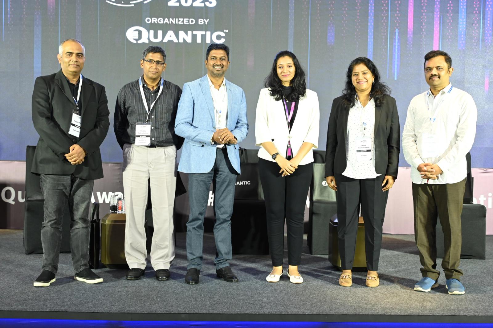 Anand Mahalingam (third from left, light blue blazer) with five fellow panelists on stage at the Quantic India DevOps panel, 2023