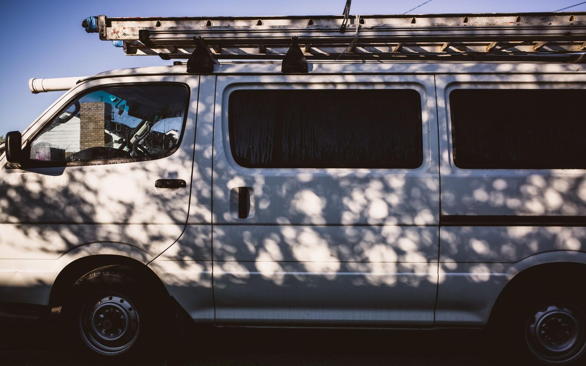 A white work van with a ladder on its roof rack, dappled with tree shadows.