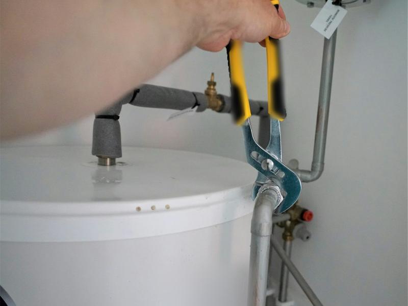 A hand using grips to tighten a pipe fitting on top of a white unvented hot water cylinder.
