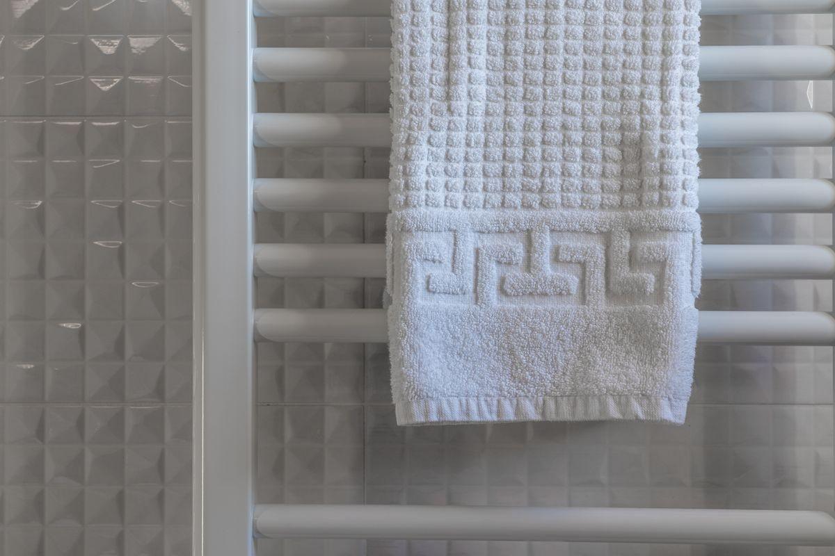 A white towel hanging on a heated towel rail against white textured tiles.
