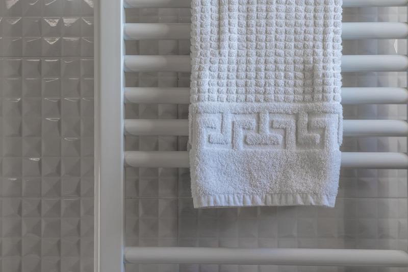 A white towel hanging on a heated towel rail against white textured tiles.