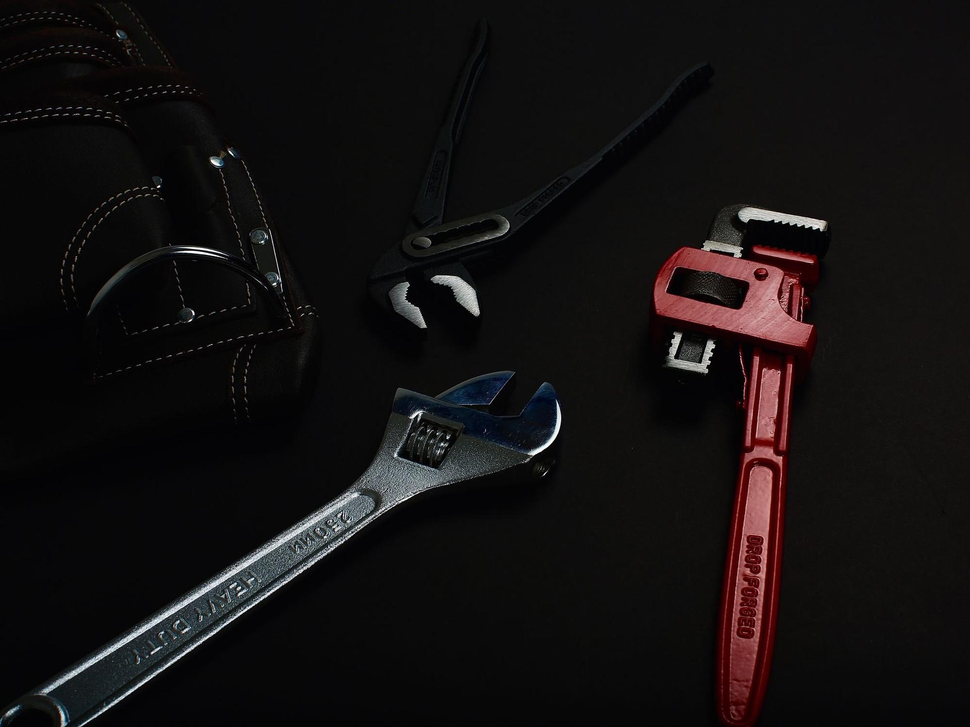 A pipe wrench, adjustable spanner, water pump pliers and a leather tool pouch on a black surface.