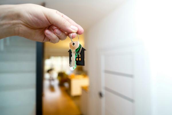 A hand holding up a set of house keys on a house-shaped keyring in a bright hallway.