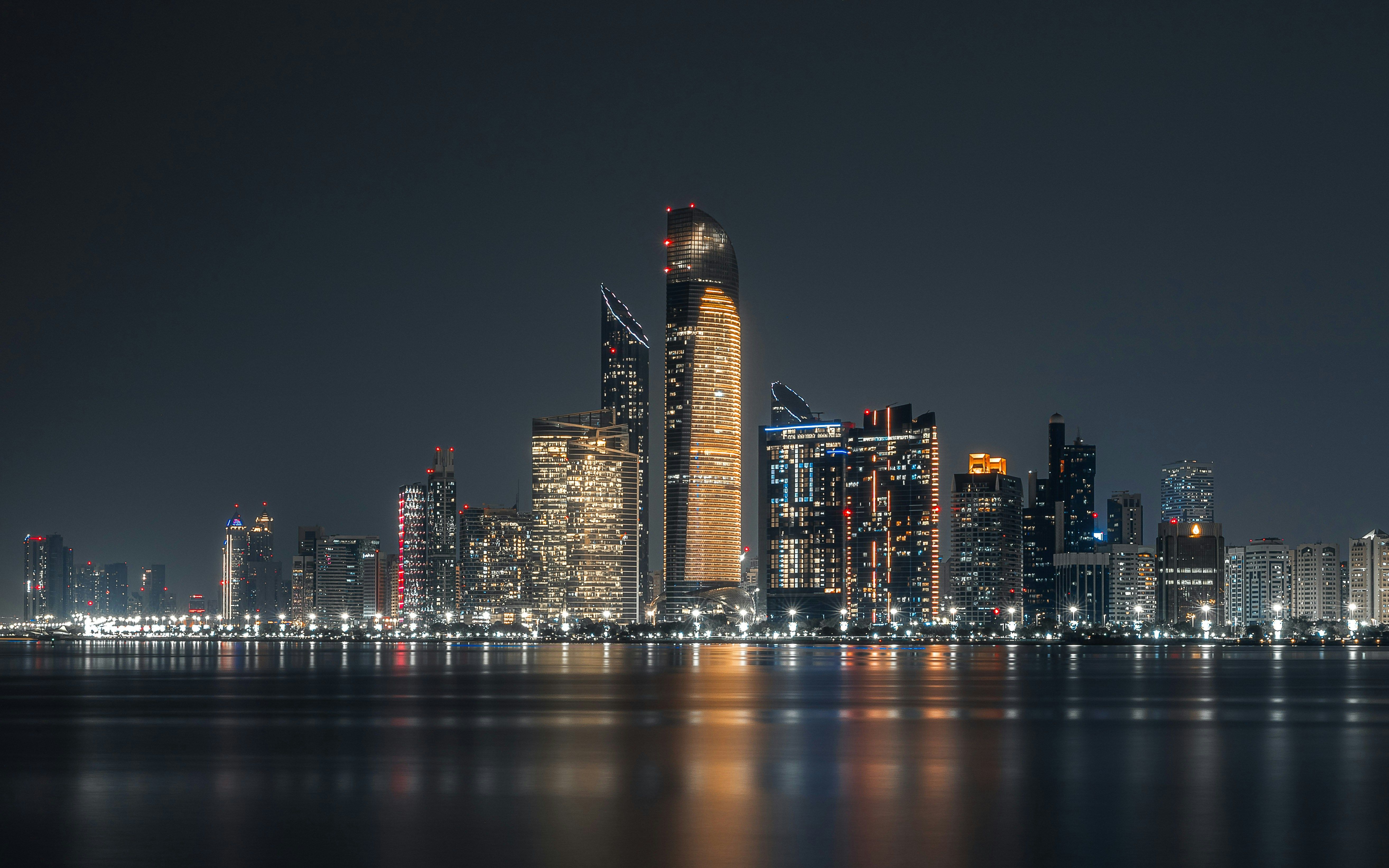 Nighttime city skyline with illuminated skyscrapers reflecting on the water.