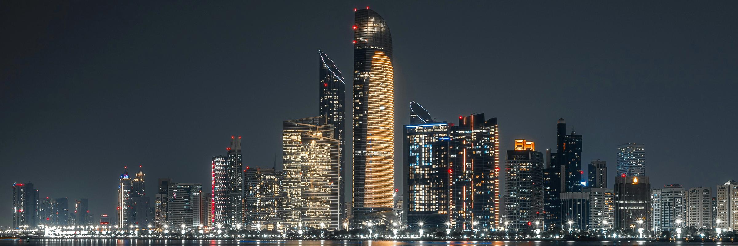 Nighttime city skyline with illuminated skyscrapers reflecting on the water.