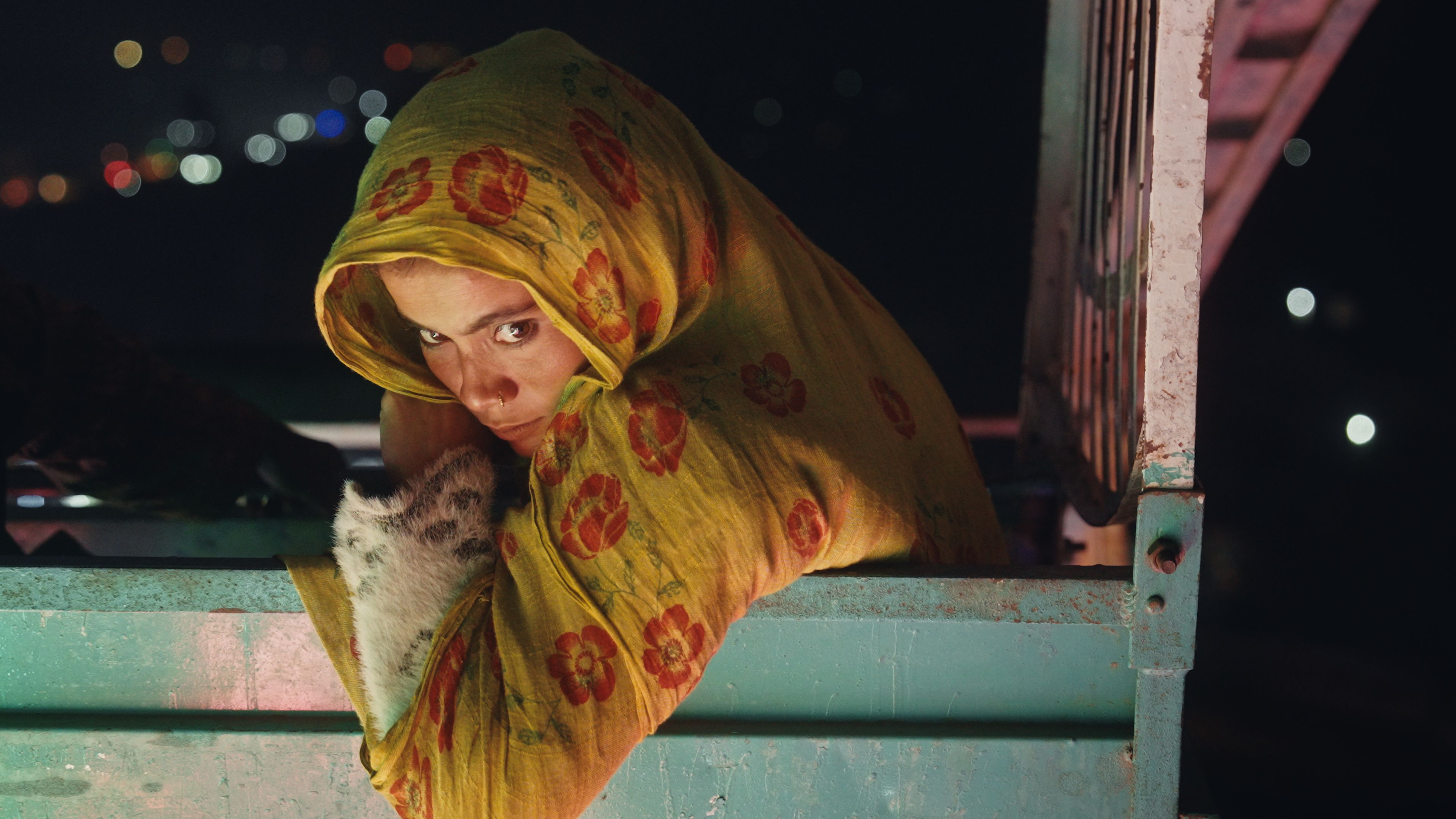 A woman in a yellow floral headscarf gazes directly, holding a spotted kitten, peeking over the edge of a metal truck bed at night.