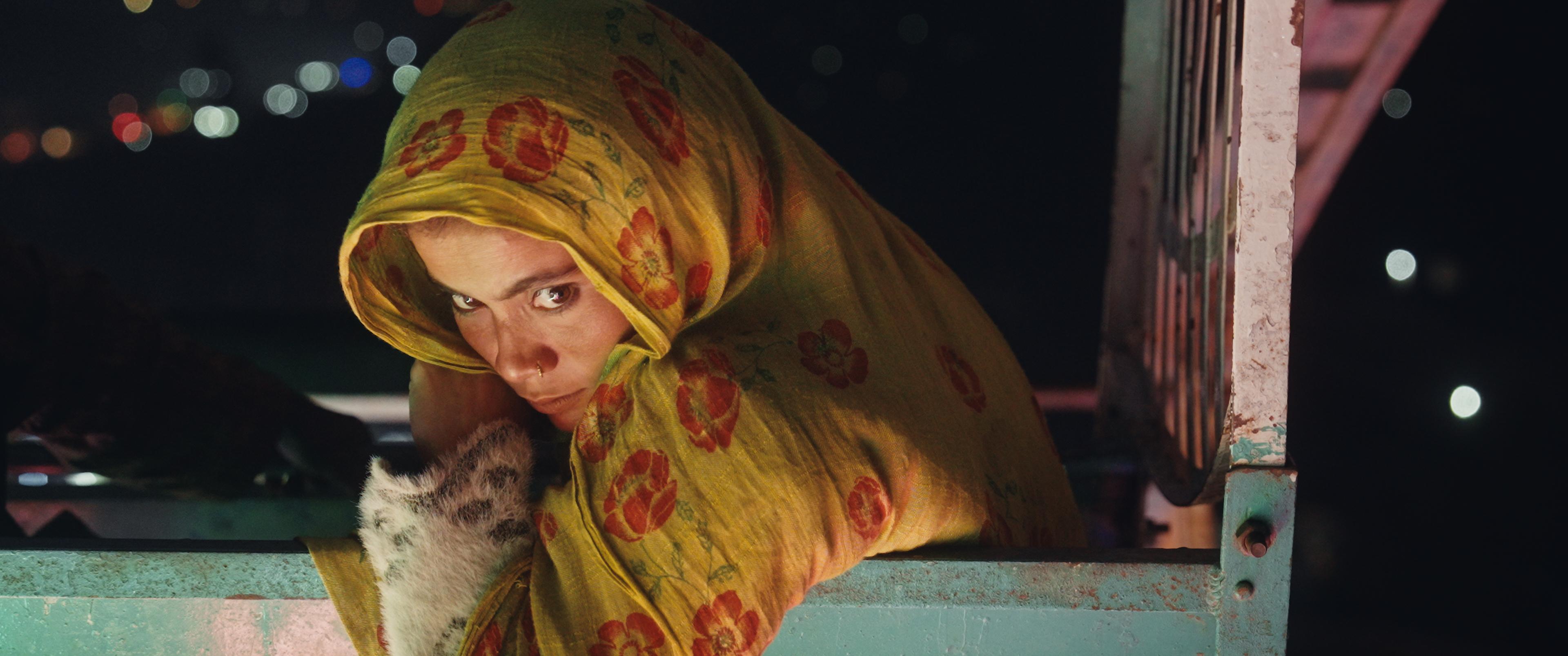 A woman in a yellow floral headscarf gazes directly, holding a spotted kitten, peeking over the edge of a metal truck bed at night.