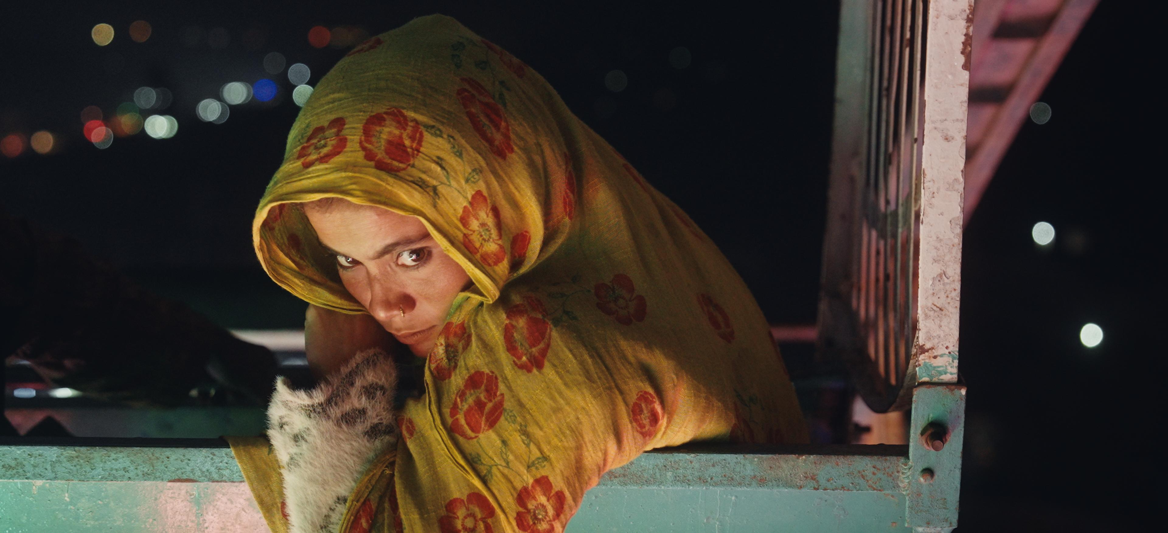 A woman in a yellow floral headscarf gazes directly, holding a spotted kitten, peeking over the edge of a metal truck bed at night.