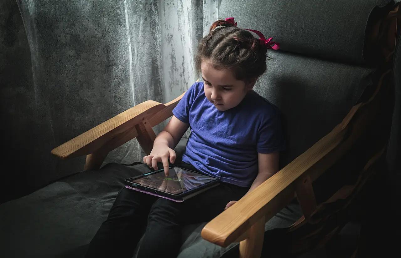 Дете на около 3–5 години седи в кресло и използва таблет до прозореца.