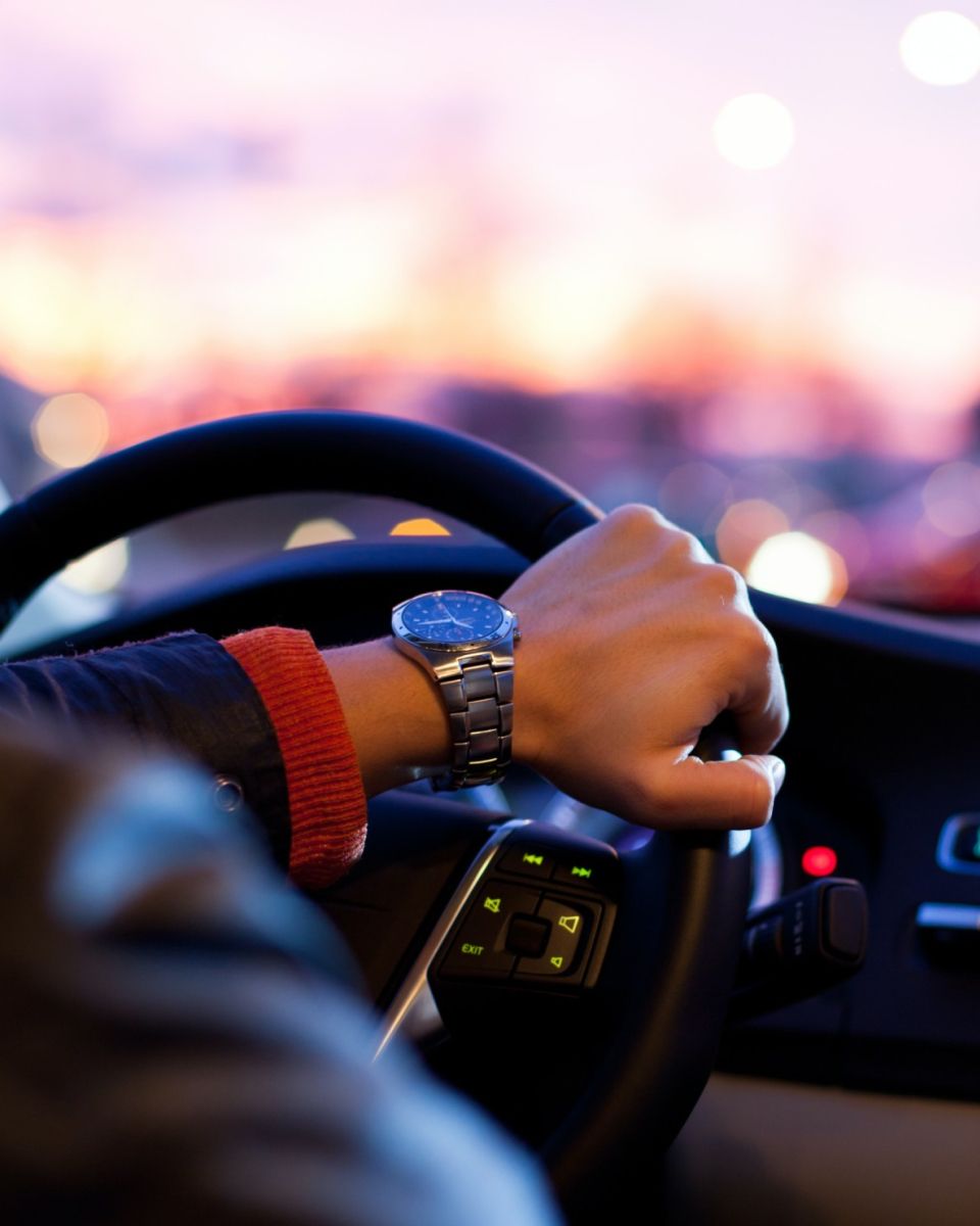 Hand wearing a wristwatch on a steering wheel with a blurred city sunset in the background.