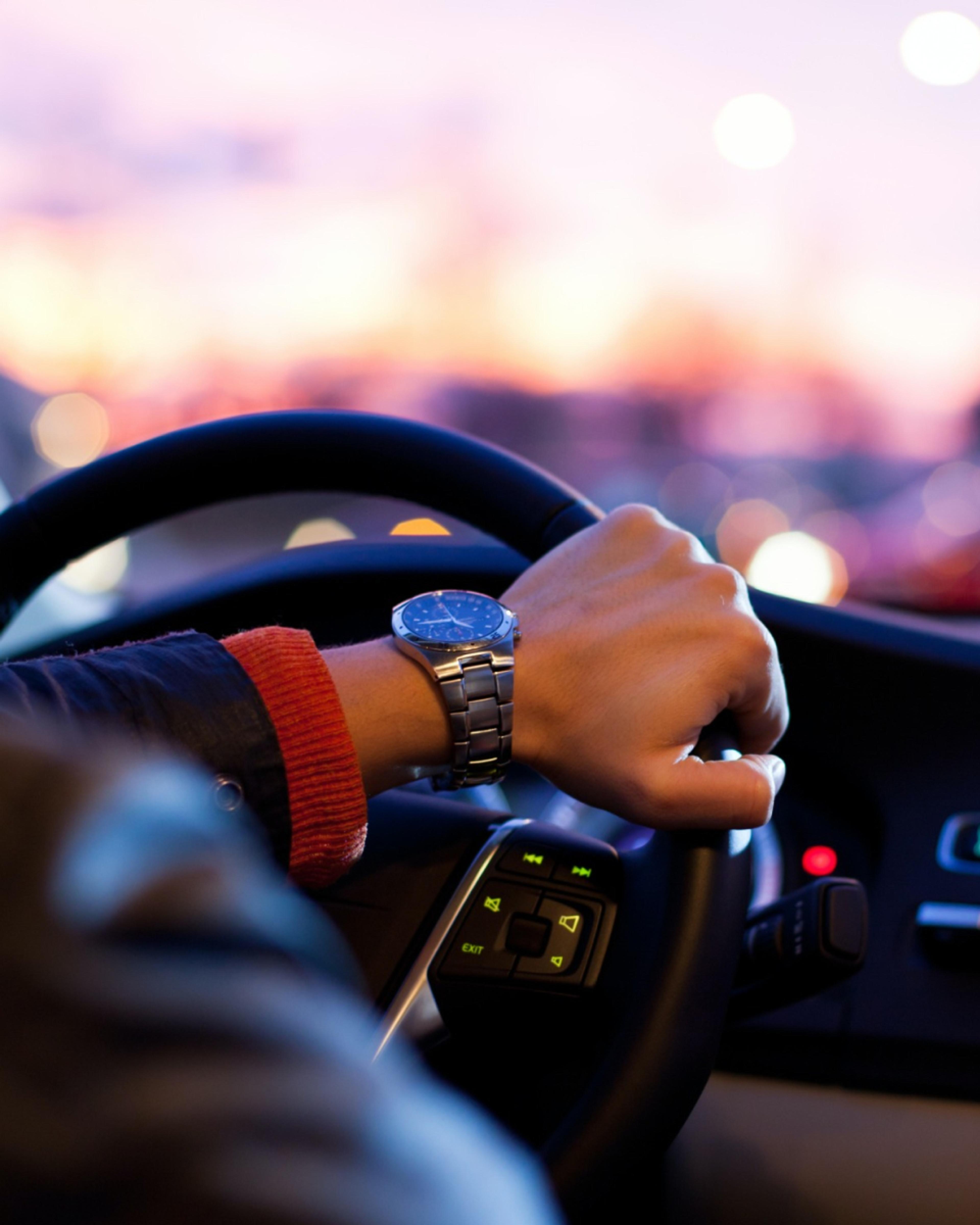 Hand wearing a wristwatch on a steering wheel with a blurred city sunset in the background.