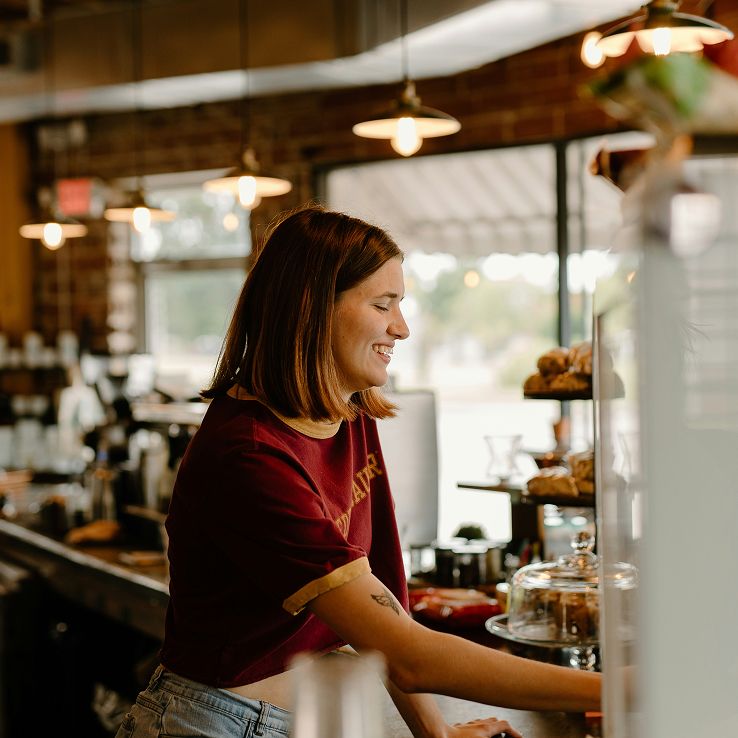 A smiling barista stands behind a cafe counter with pastries in the background.