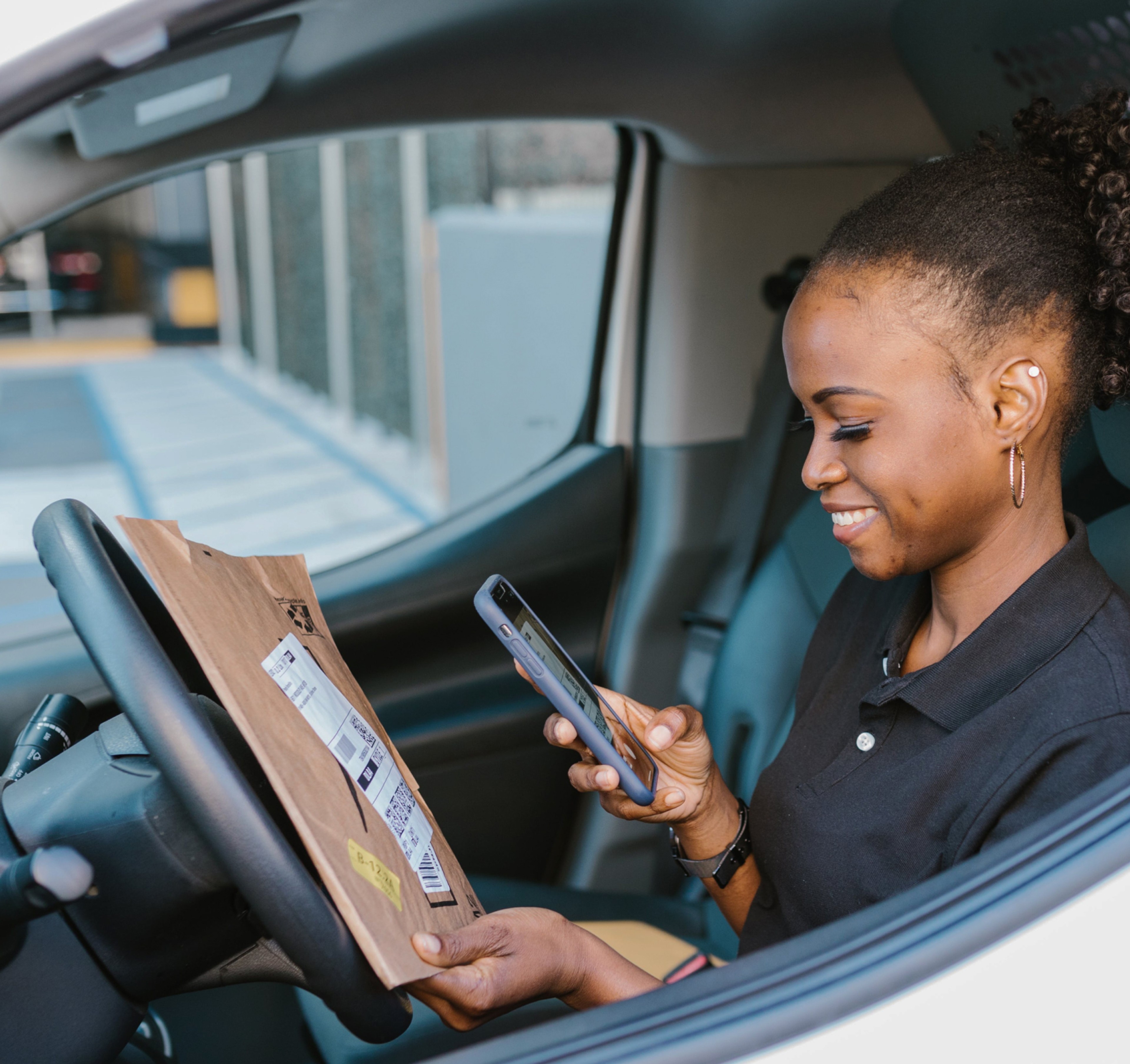 Smiling delivery driver sitting in a vehicle, scanning a package with a smartphone, representing a stress-free delivery experience.