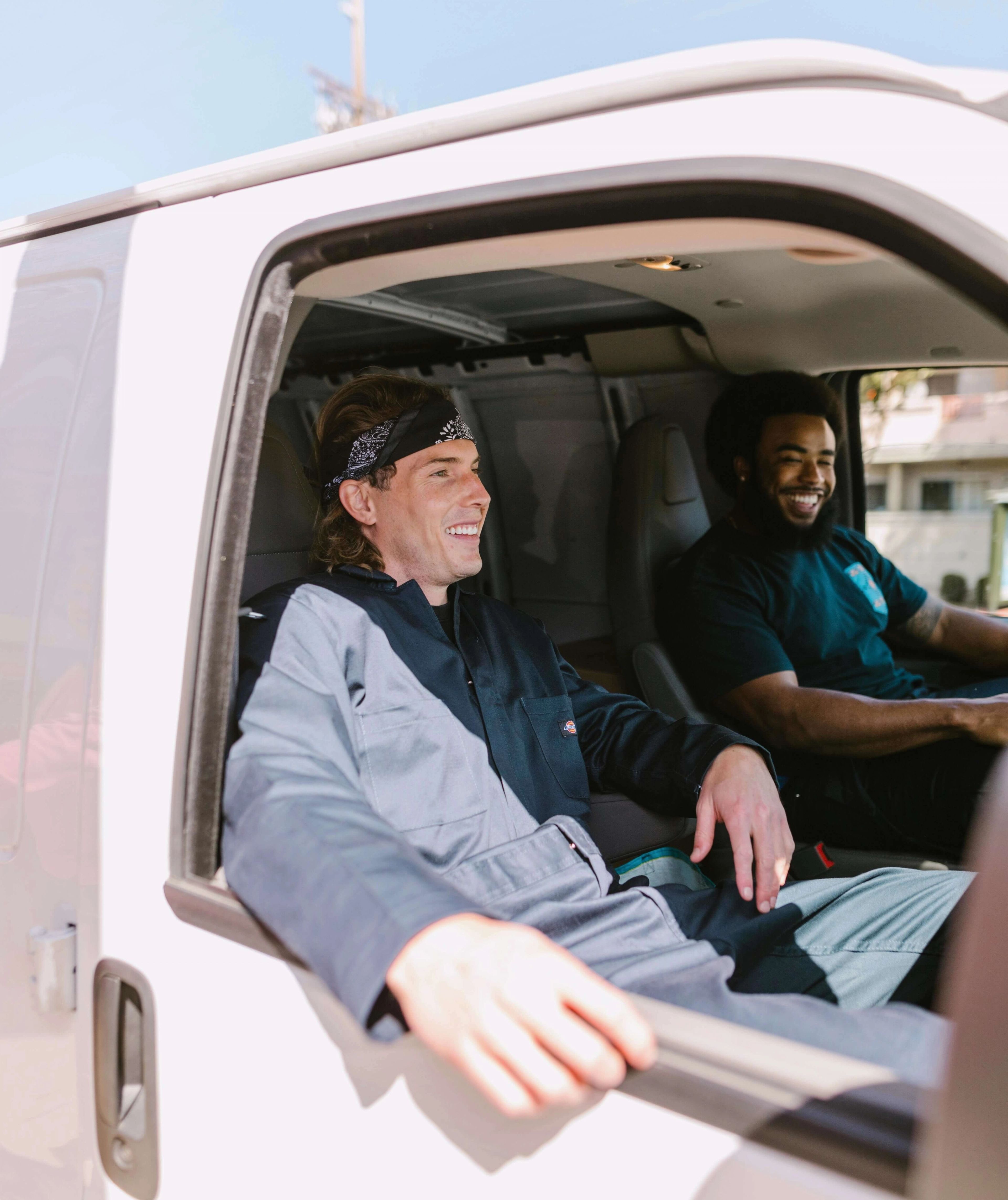 Two delivery drivers sitting side by side in the cab of a white van, smiling and chatting.