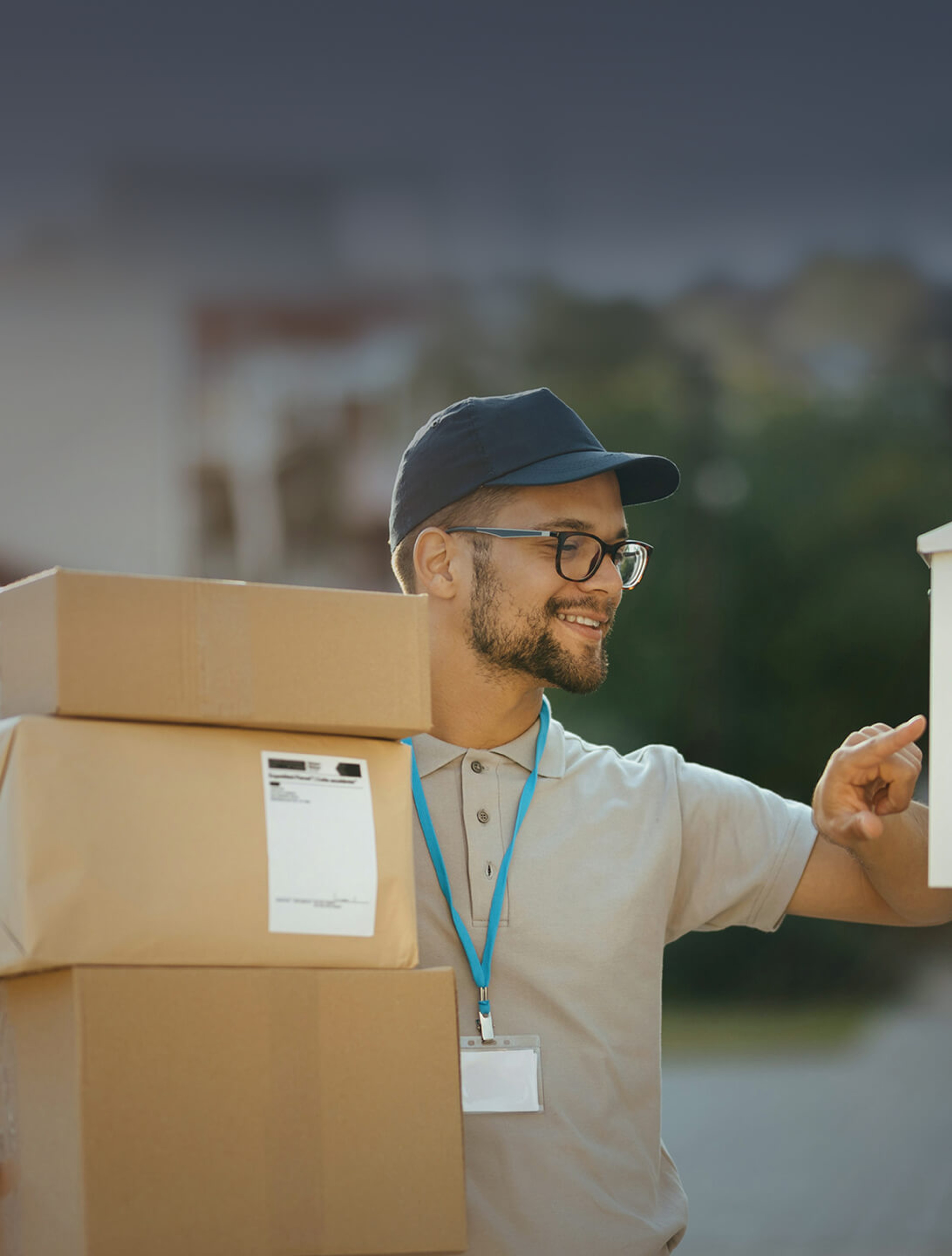 Smiling delivery driver wearing a cap and glasses, holding three cardboard boxes and pointing toward a building.