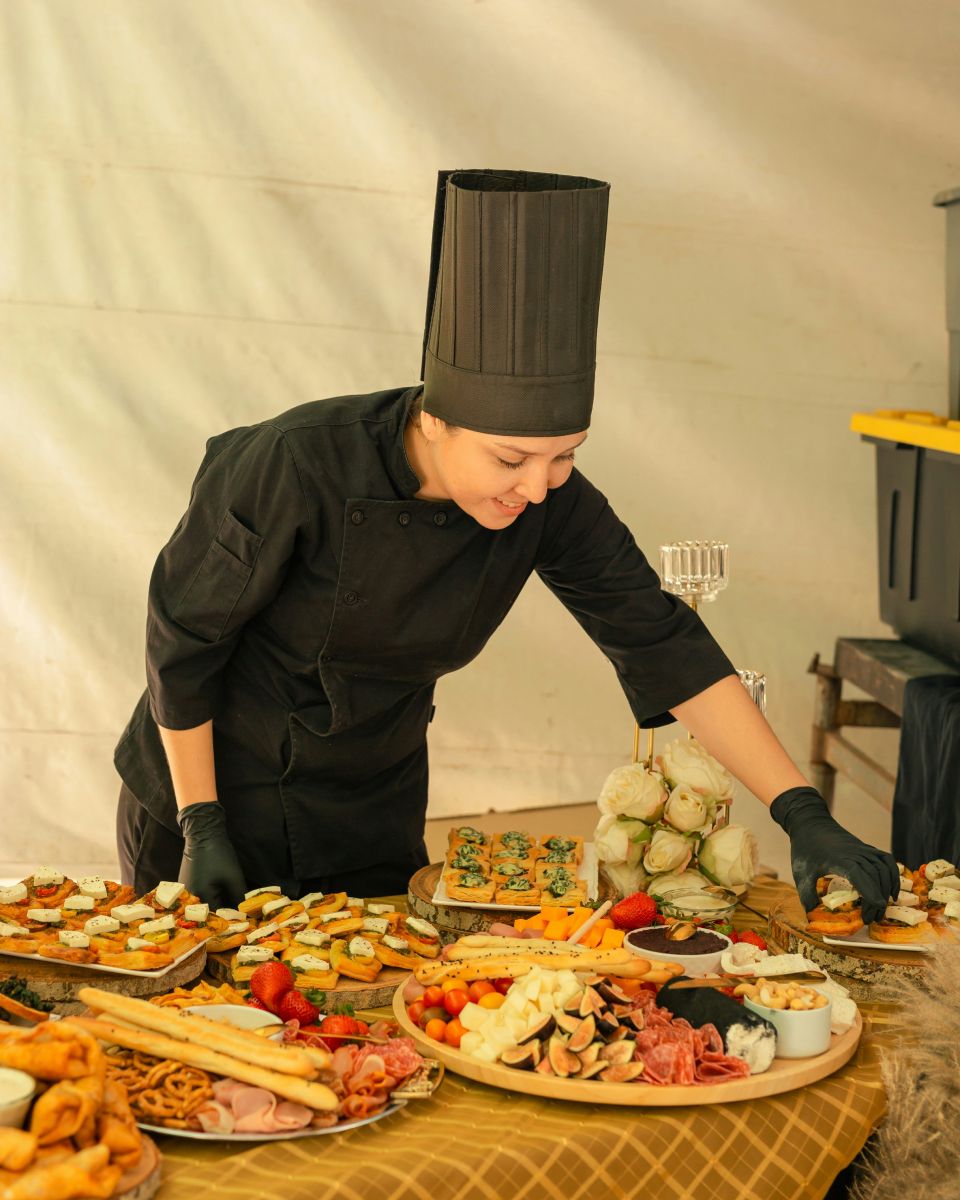 A smiling chef in a black uniform and hat adjusts food on a table laden with appetizers and charcuterie.