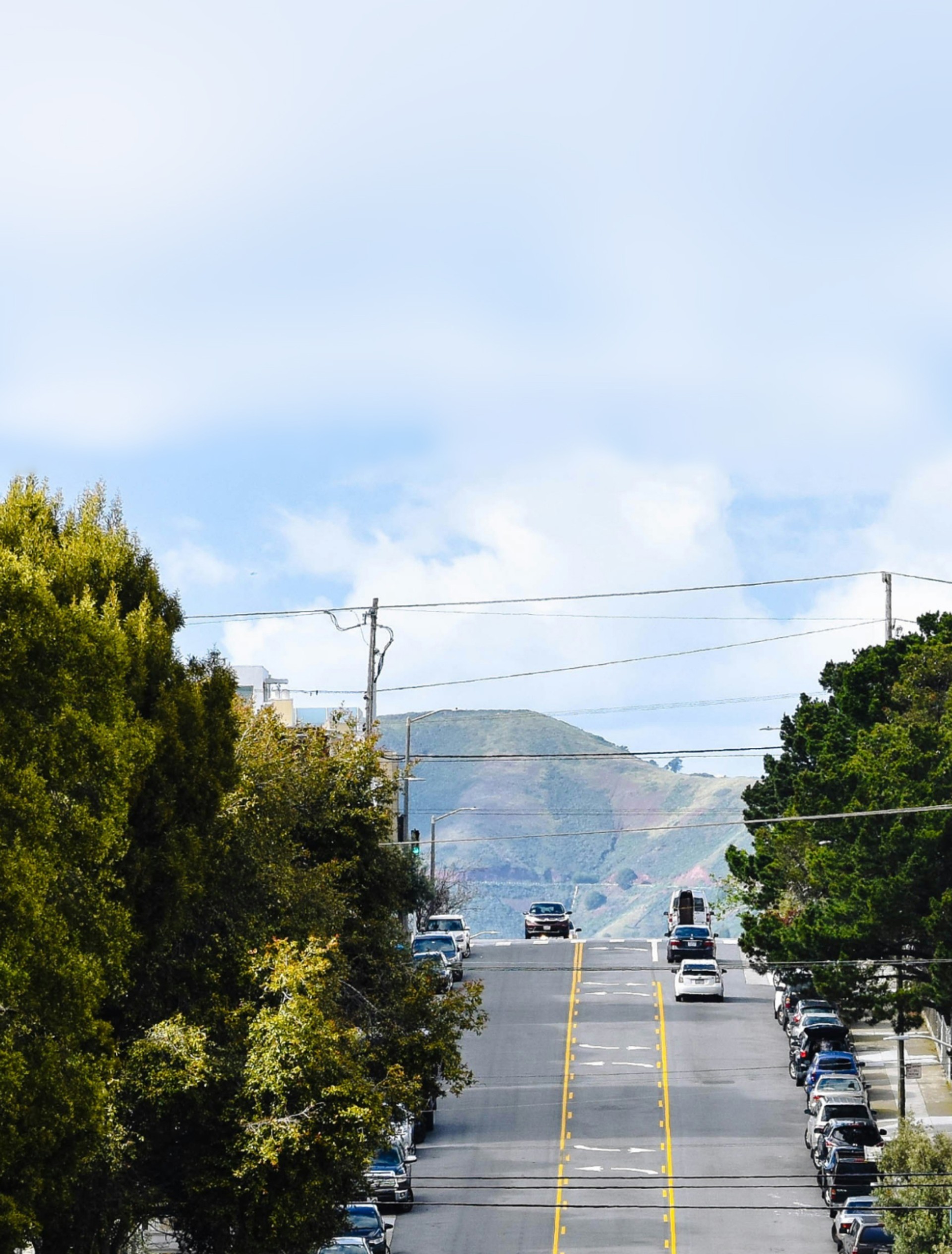 A steep street lined with trees and parked cars leads up to a distant mountain under a cloudy sky.