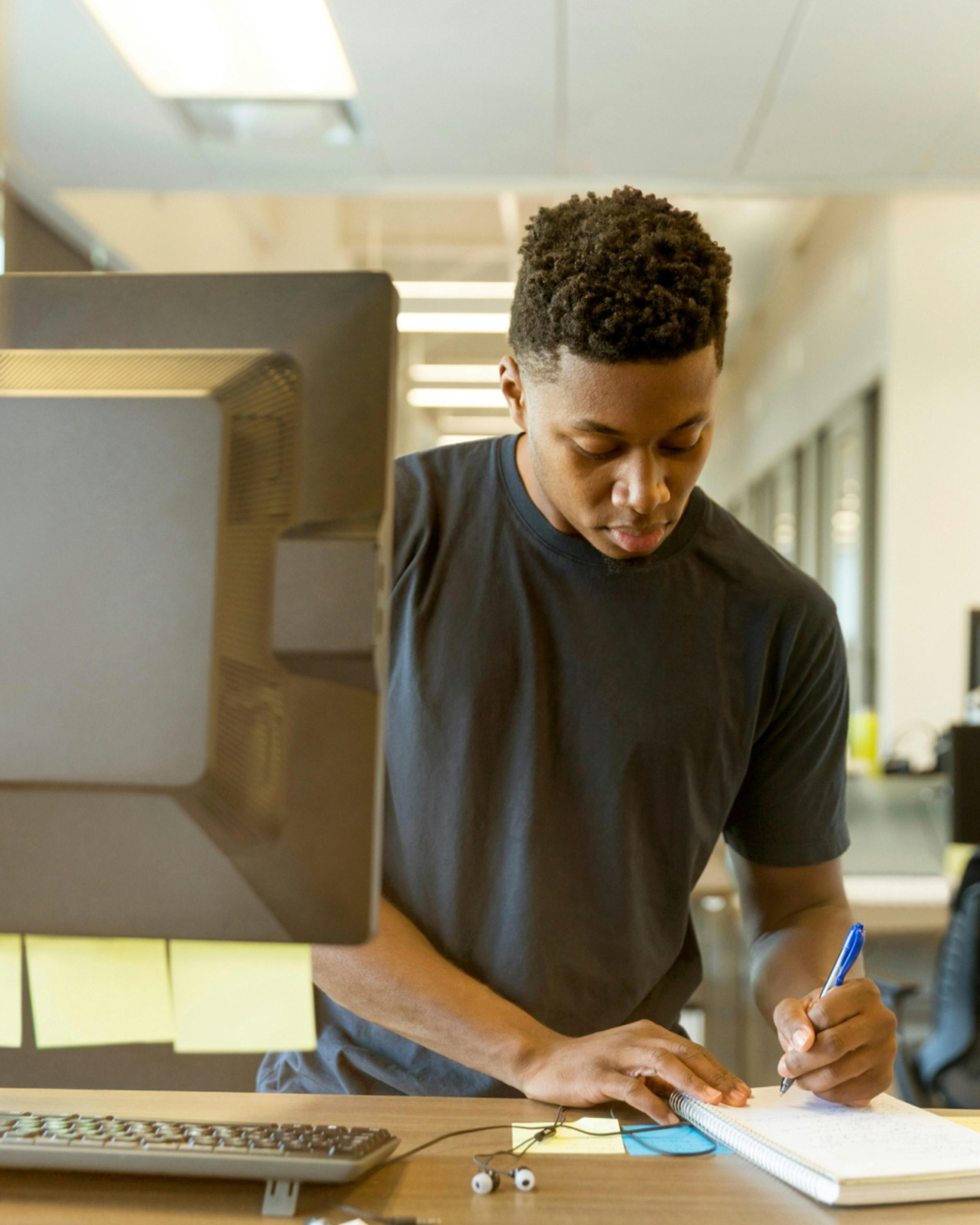 A young Black man stands at a desk, writing in a spiral notebook with a pen.