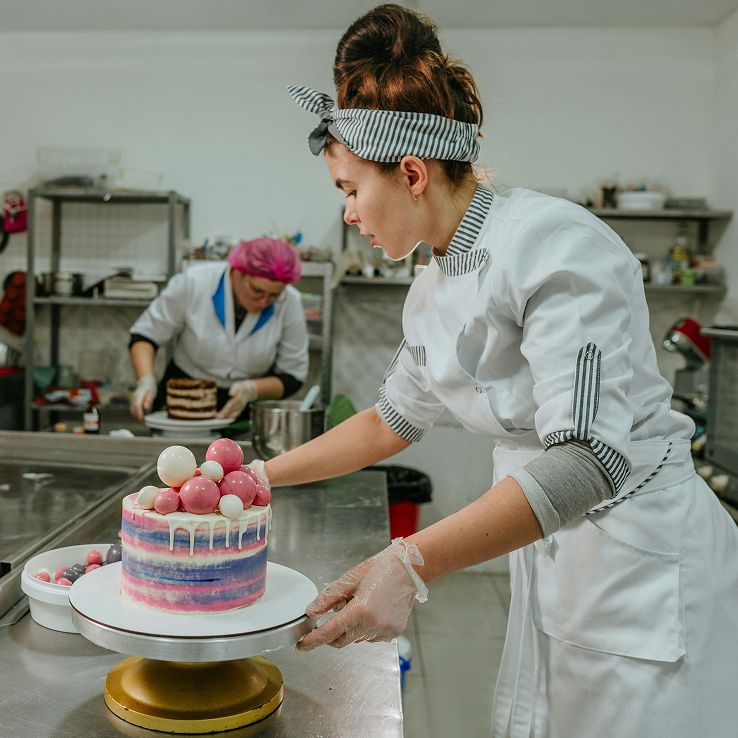 A baker in a white coat decorates a colorful cake topped with spheres, another baker works in the background.