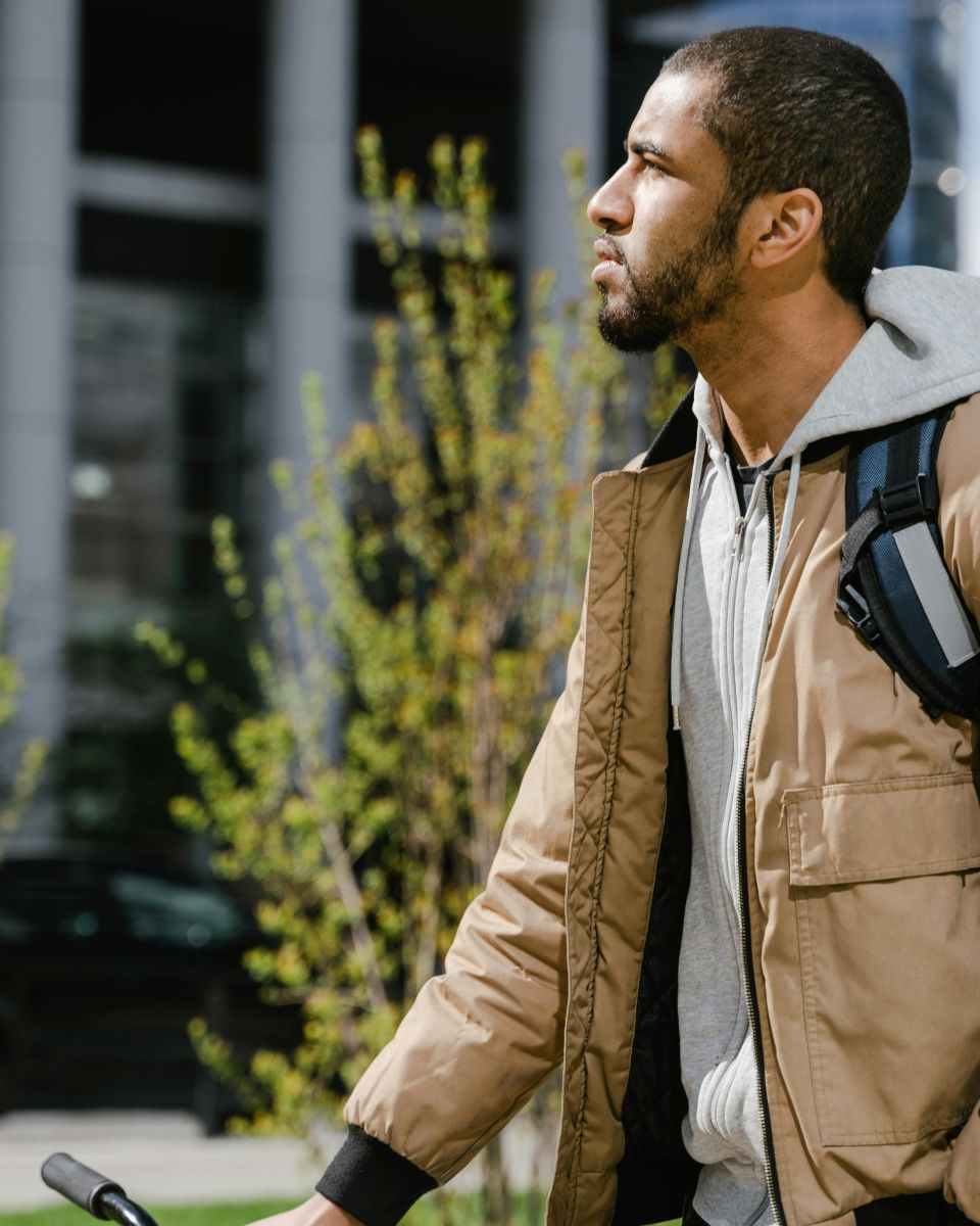 Profile of a young man with a beard, wearing a tan jacket and a backpack, looking right outdoors.