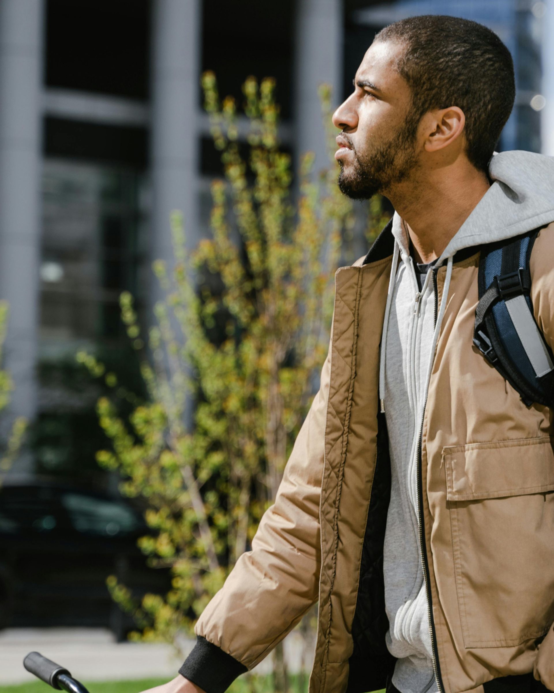 Profile of a young man with a beard, wearing a tan jacket and a backpack, looking right outdoors.