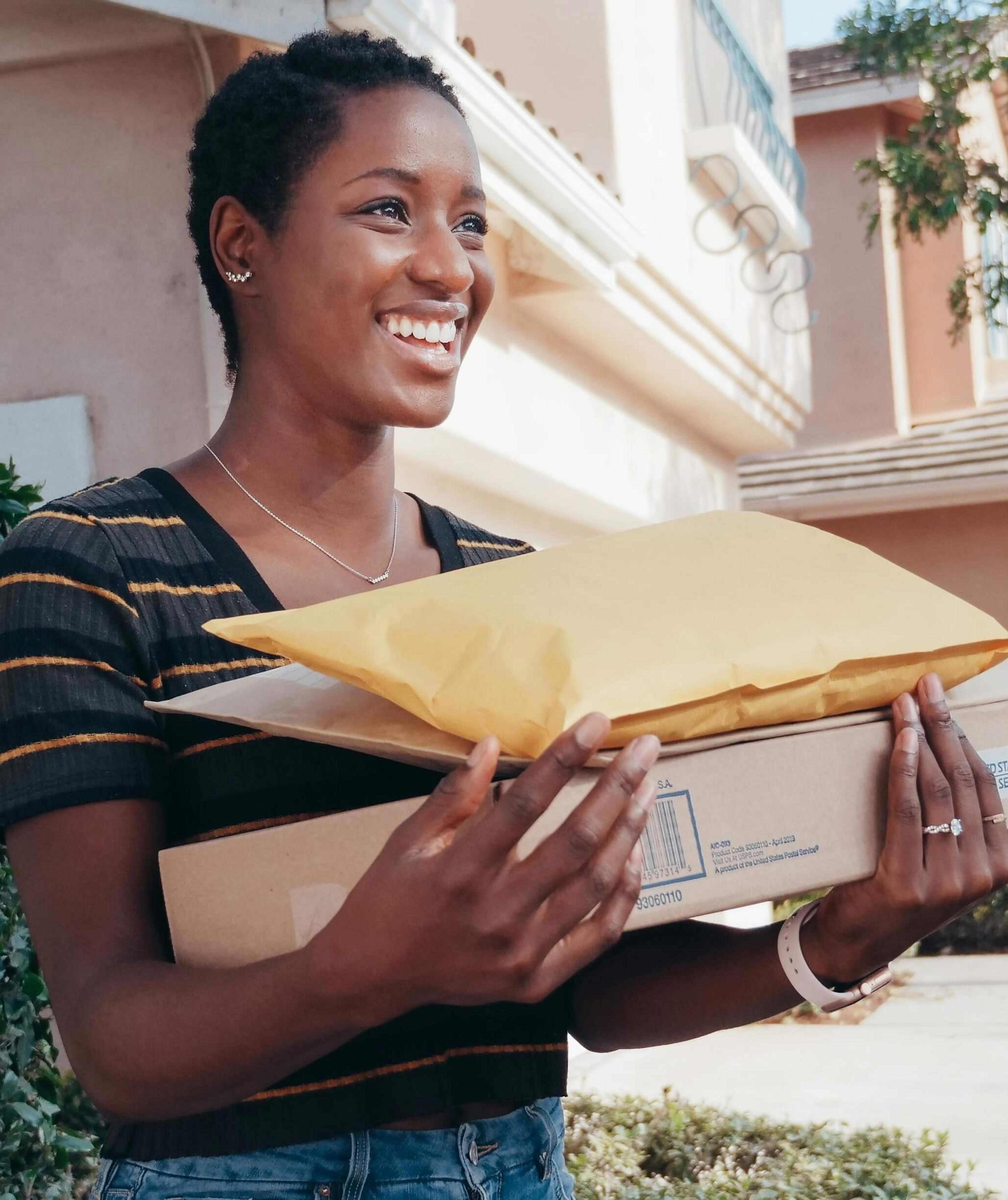 Smiling woman standing outside holding multiple delivered packages, illustrating a positive delivery experience and recipient satisfaction in a residential setting.