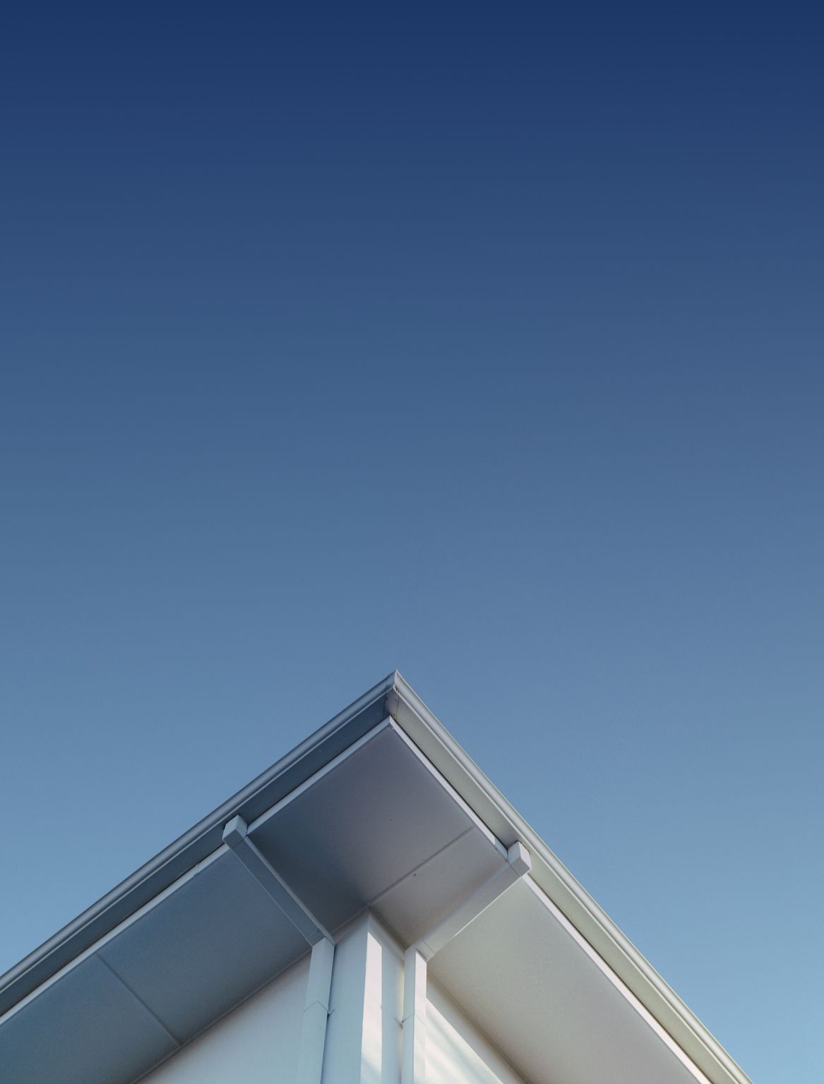 A sharp corner of a white building's roof and gutters against a clear blue sky.