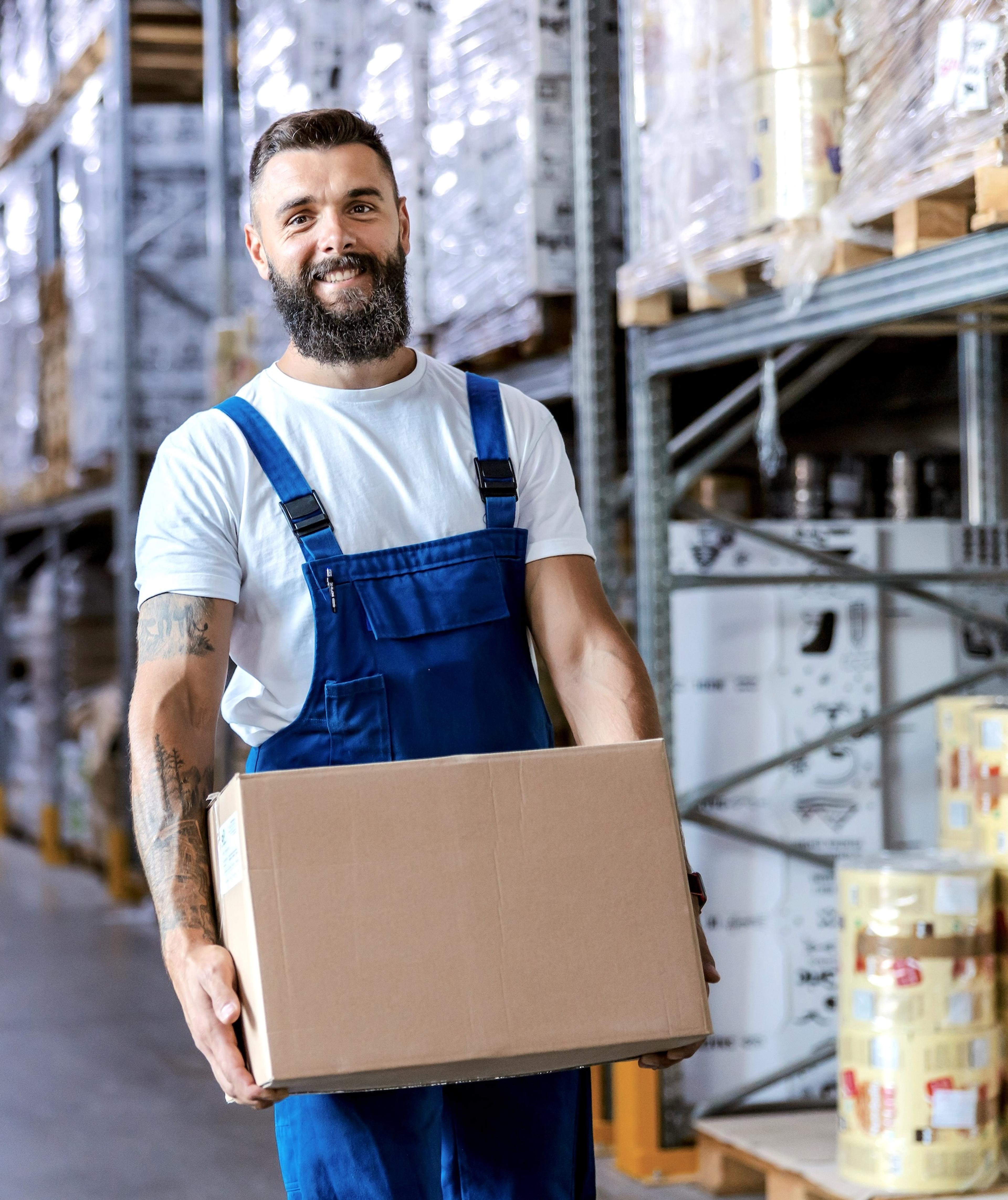 A bearded man in a white T-shirt and blue overalls smiling as he carries a large cardboard box down a warehouse aisle lined with tall metal shelving stacked with wrapped pallets and boxes.