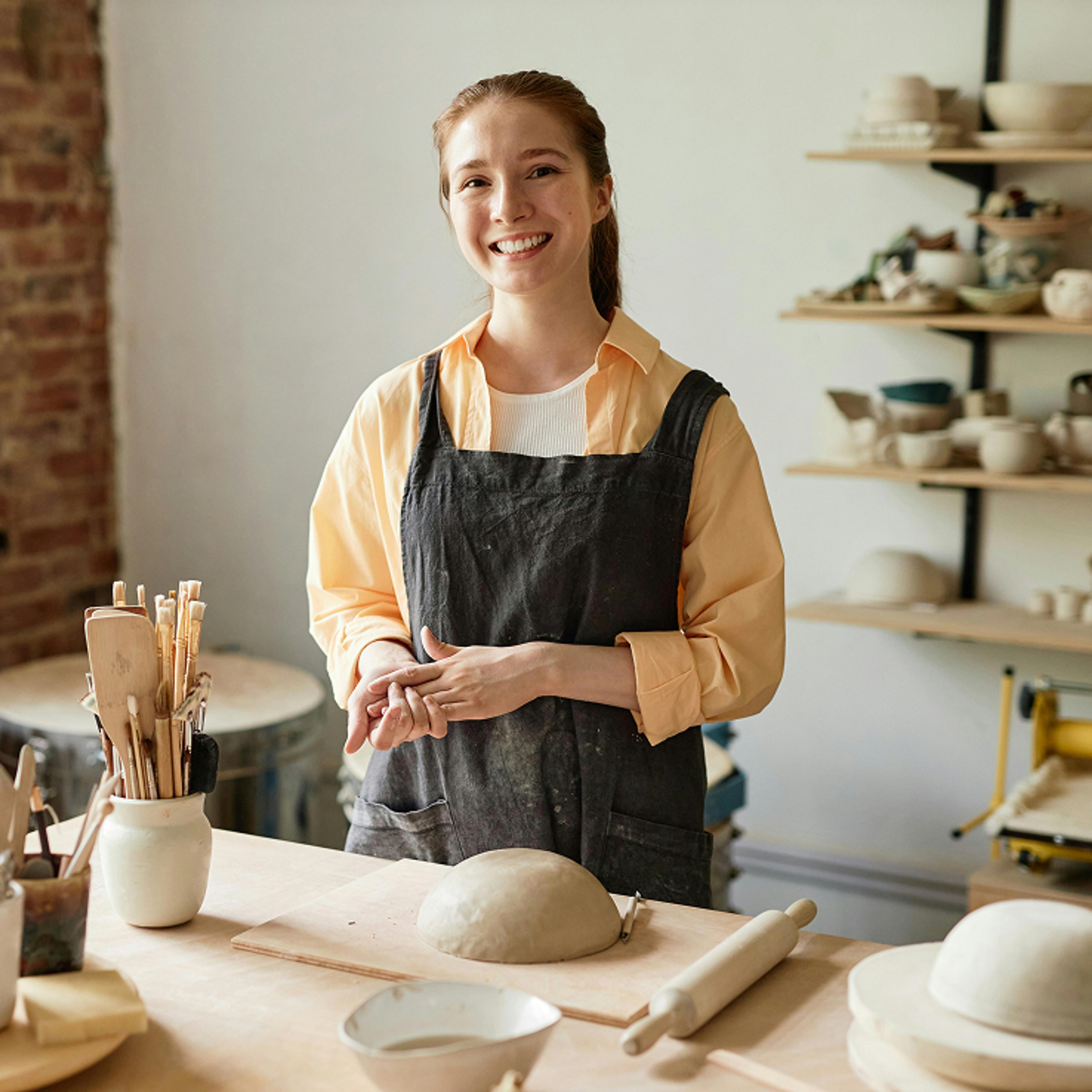 A smiling young woman in an apron stands at a pottery workbench with clay and tools.