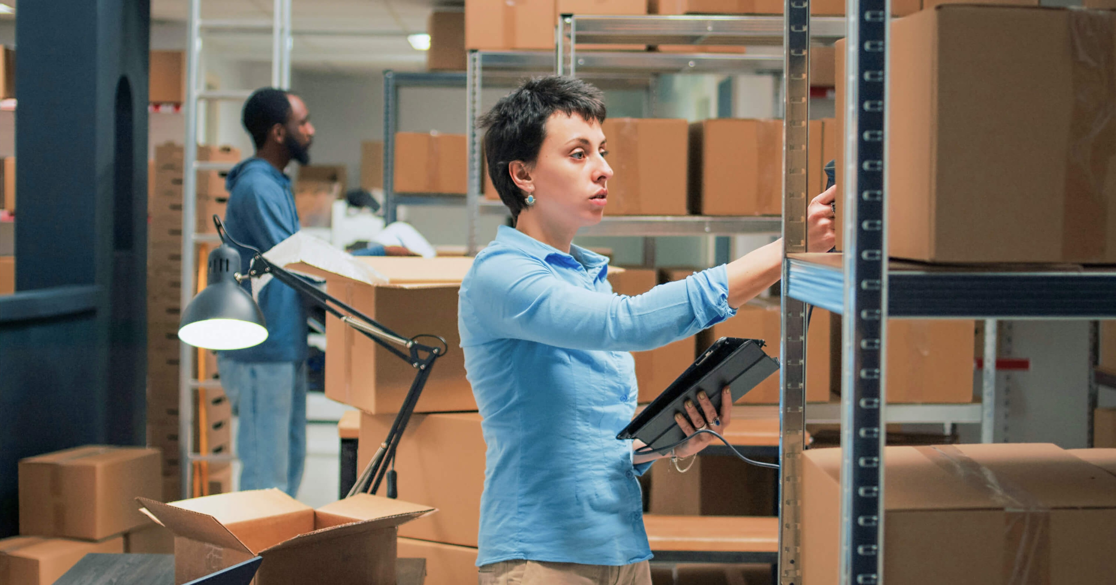 Warehouse worker scanning packages and using a tablet while organizing inventory on metal shelves, with cardboard boxes stacked in the background.