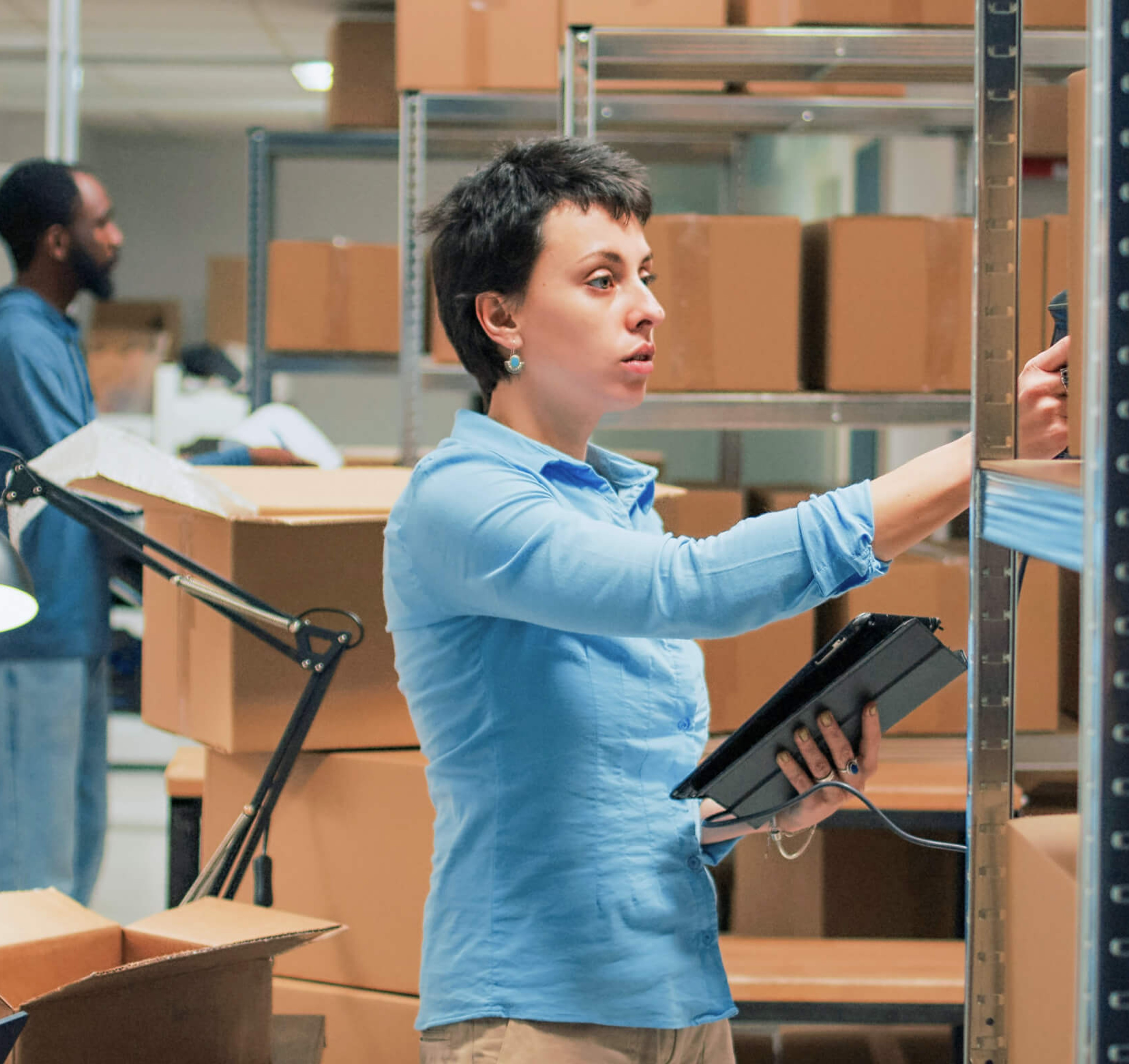 Warehouse worker scanning packages and using a tablet while organizing inventory on metal shelves, with cardboard boxes stacked in the background.