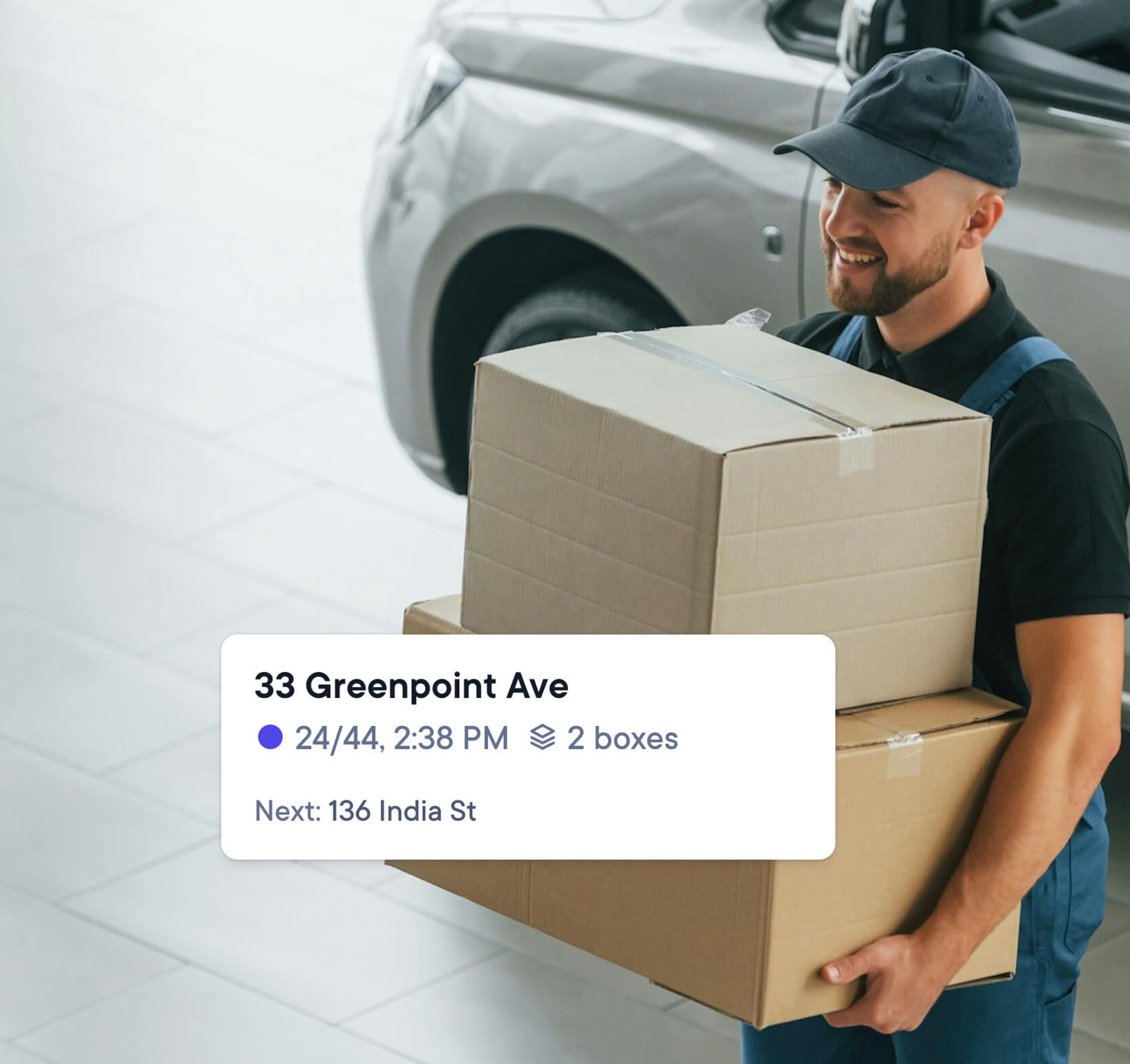 Delivery driver in uniform carrying two large cardboard boxes away from a van, with a location tag showing ‘33 Greenpoint Ave’ and details for the next delivery stop at ‘136 India St’