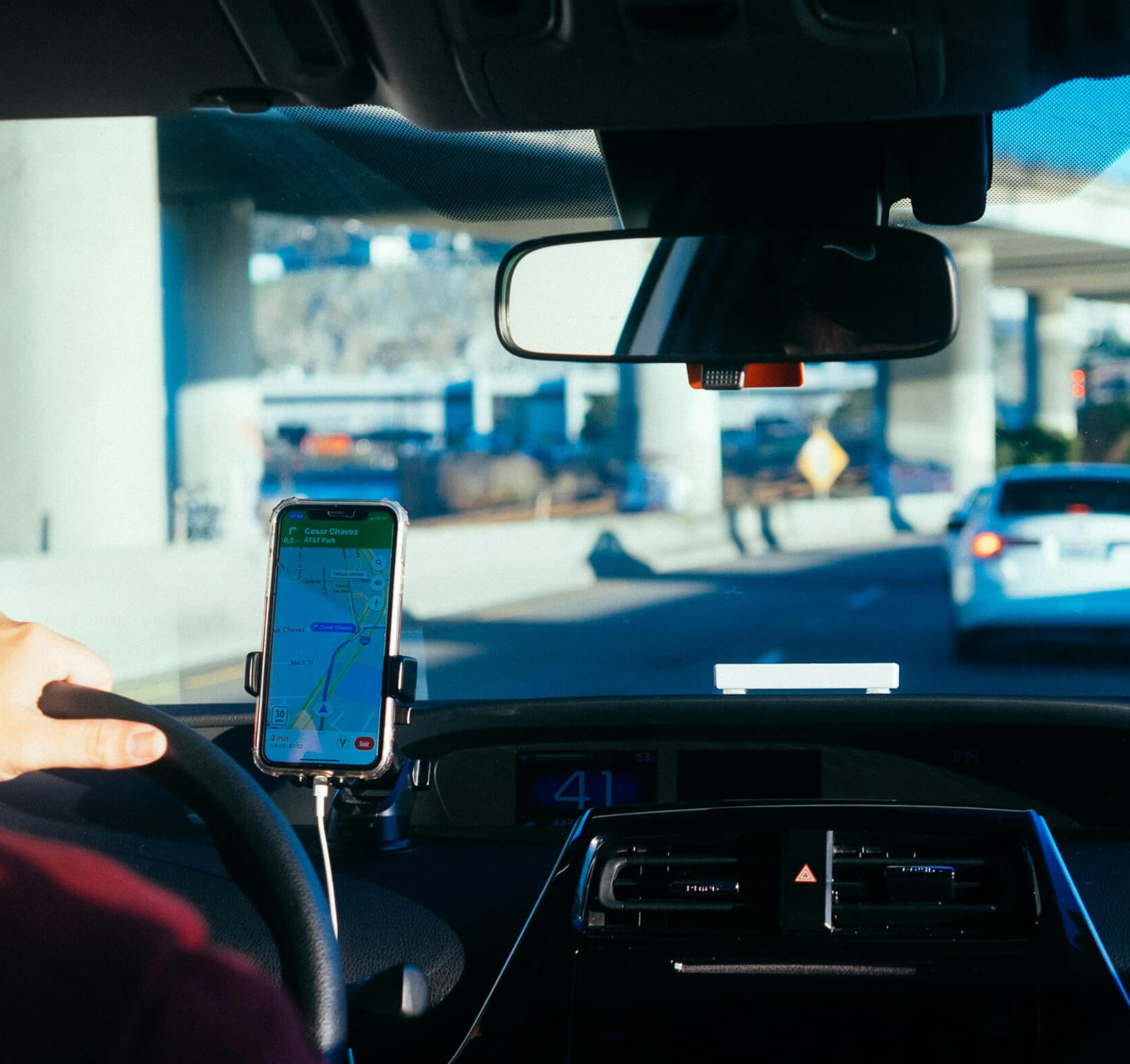 Driver navigating city streets using a smartphone-mounted GPS system, viewed from inside the car with a highway and overpasses ahead.