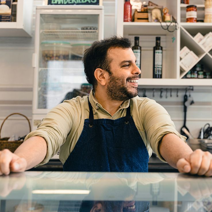A smiling man with a beard and dark apron stands behind a counter.