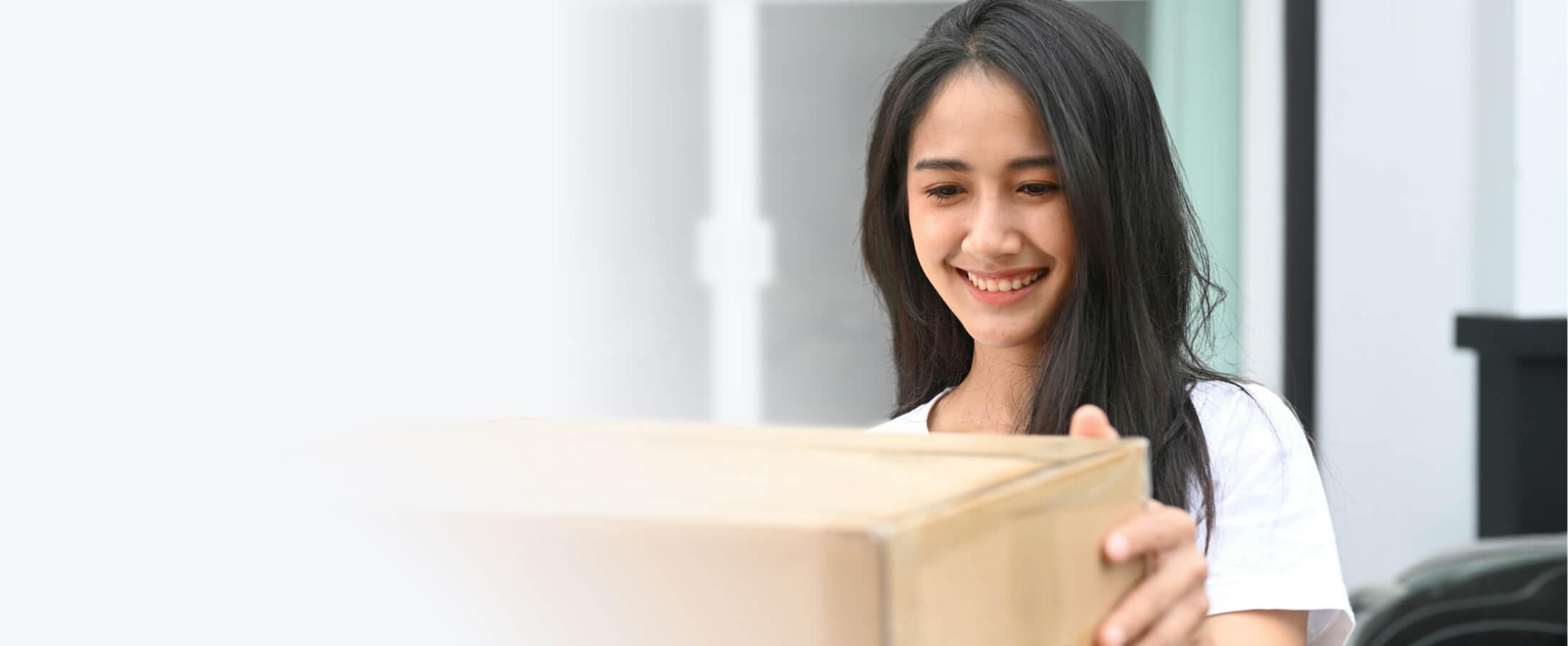 A young woman smiling as she holds a large cardboard box in front of a blurred doorway.