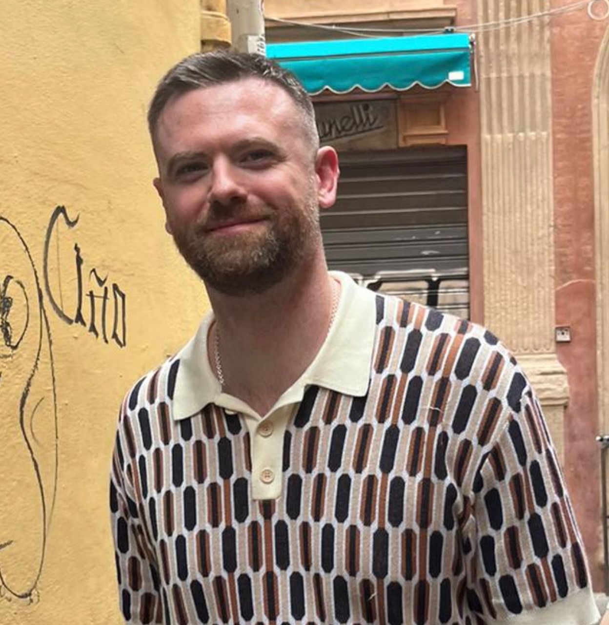 A smiling man with a beard and patterned polo shirt stands in front of a yellow wall with graffiti, with a blurred street scene behind him.