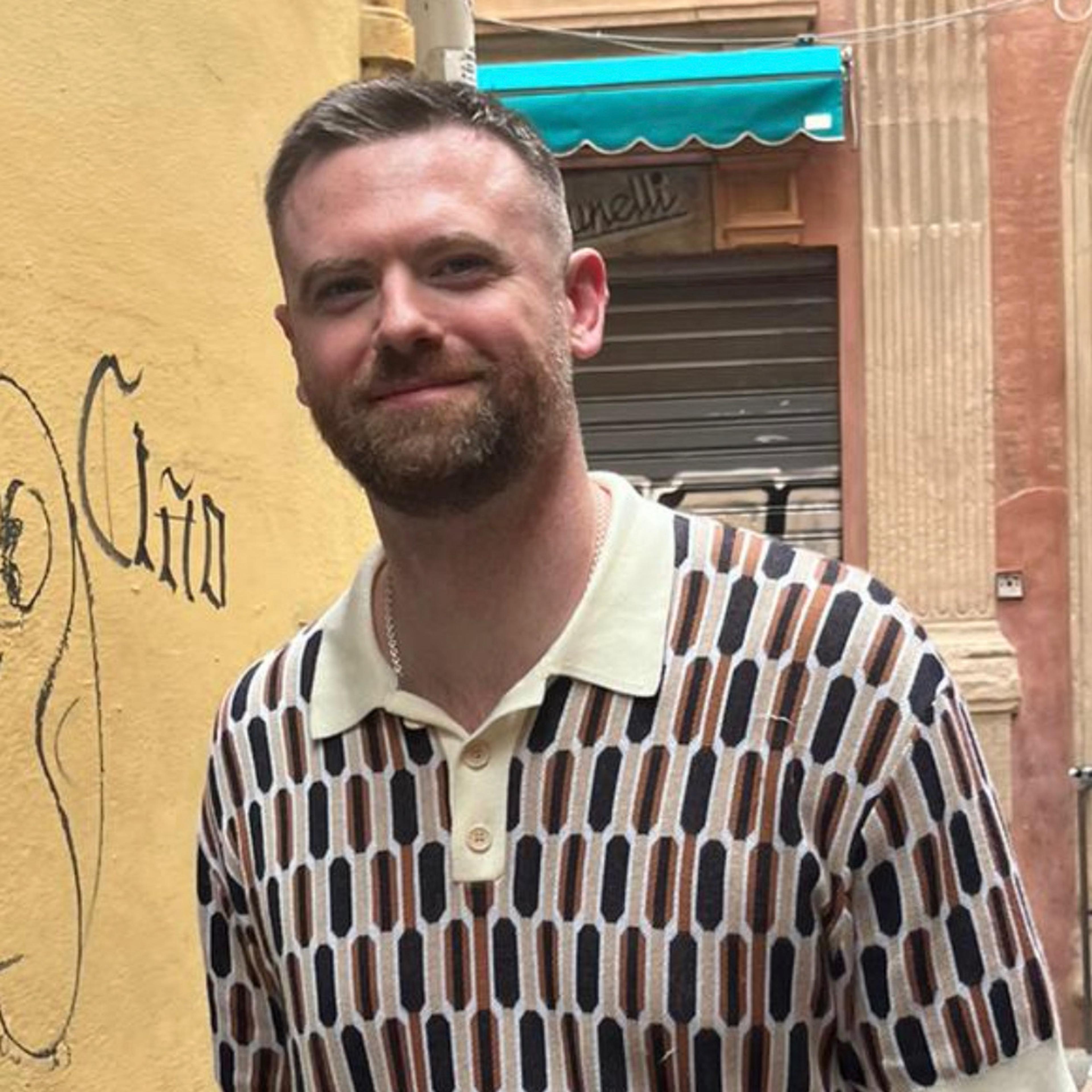 A smiling man with a beard and patterned polo shirt stands in front of a yellow wall with graffiti, with a blurred street scene behind him.