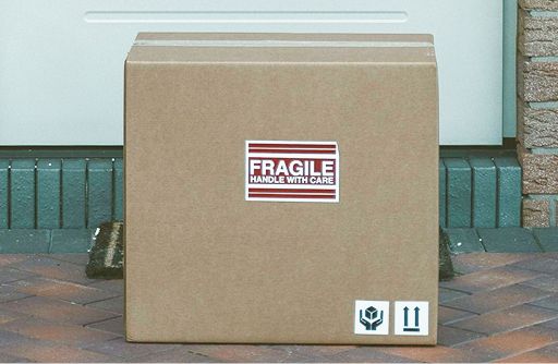 A brown cardboard box with a ‘FRAGILE HANDLE WITH CARE’ sticker and shipping symbols sits on a doorstep.