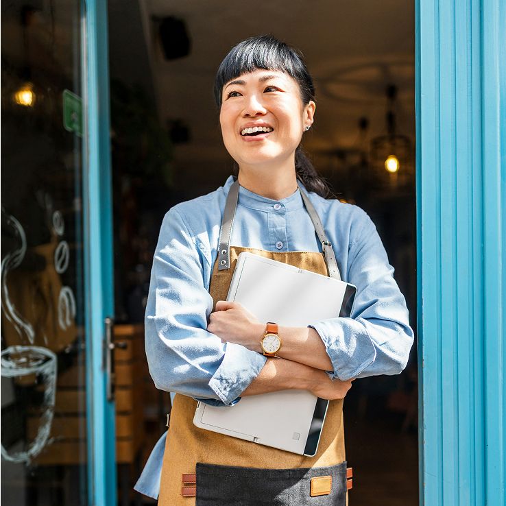Smiling Asian woman in an apron holds a tablet, standing in a shop doorway.