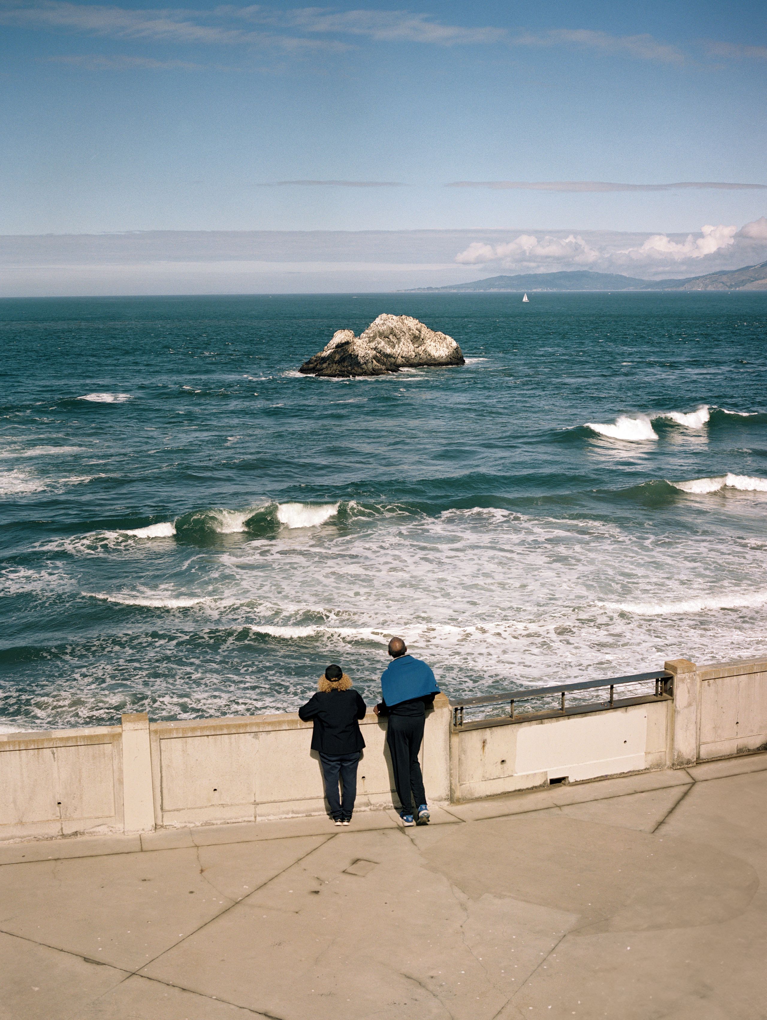 Two figures at the seawall above Ocean Beach, Seal Rocks offshore beneath a wide, bright sky