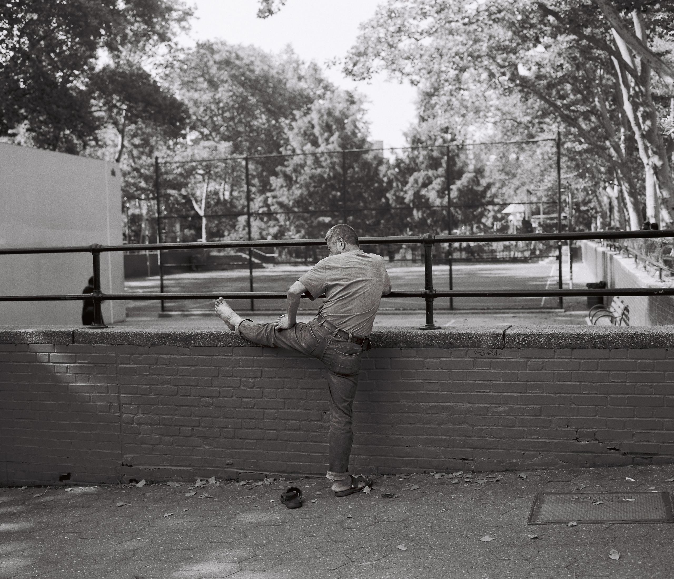 An older man stretches one leg along a park railing in morning light, black-and-white film
