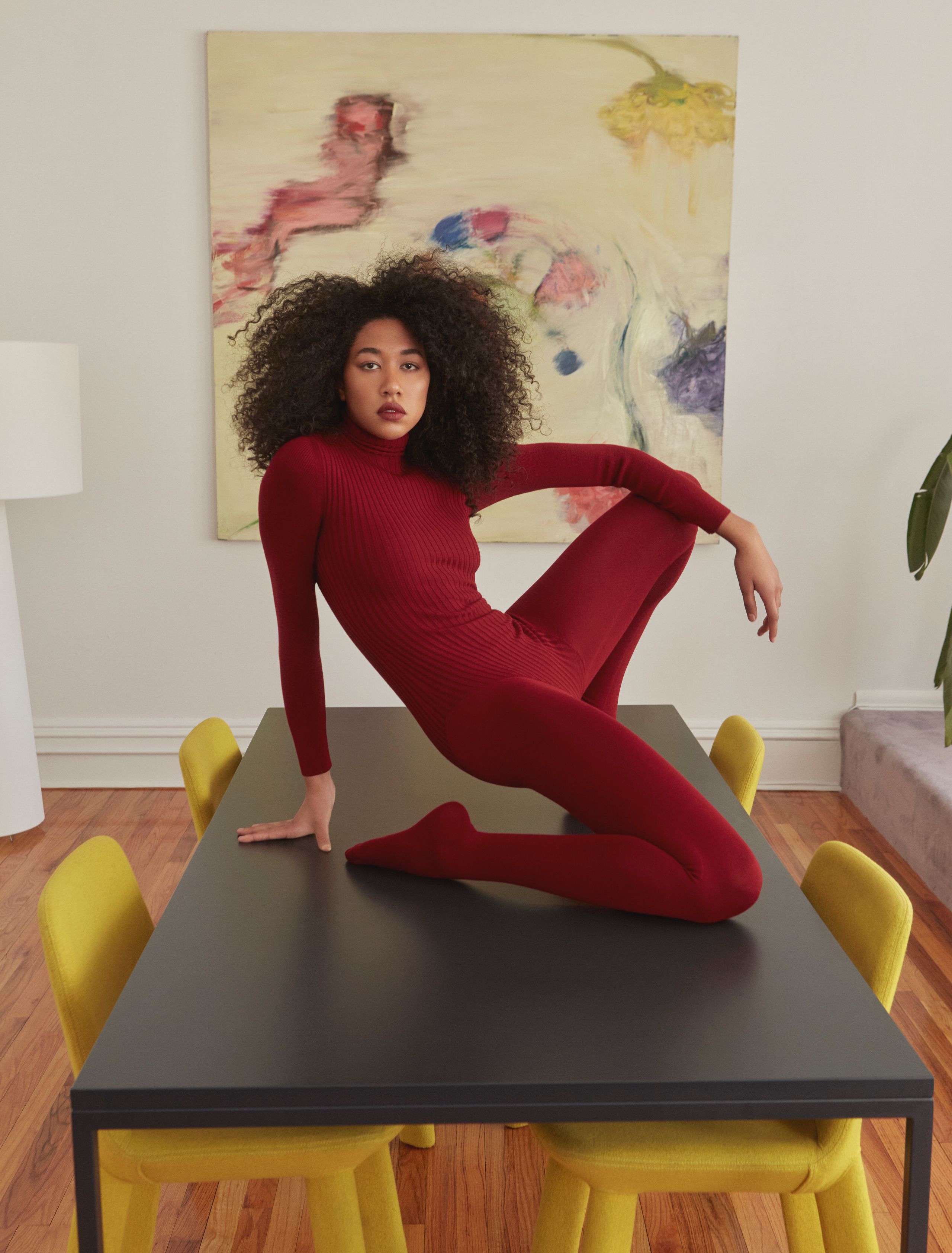 Aoki Lee Simmons seated on a dark table in red knit, leaning back on one hand with legs folded, gaze to camera