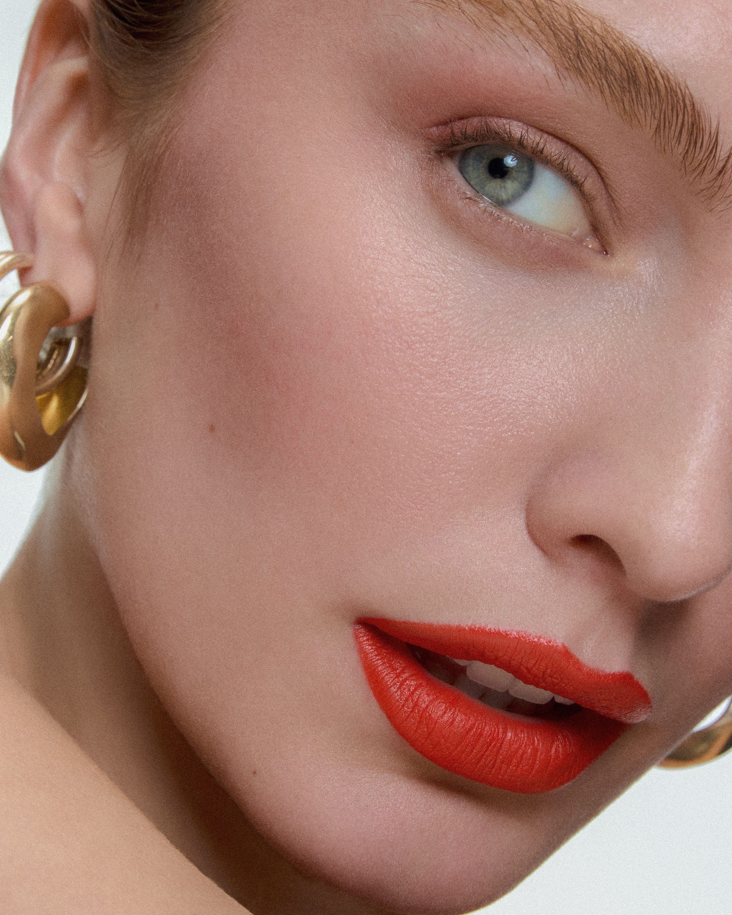 Close-up of coral lips, brushed-up brows and a chunky gold AGMES hoop, head tilted to the side