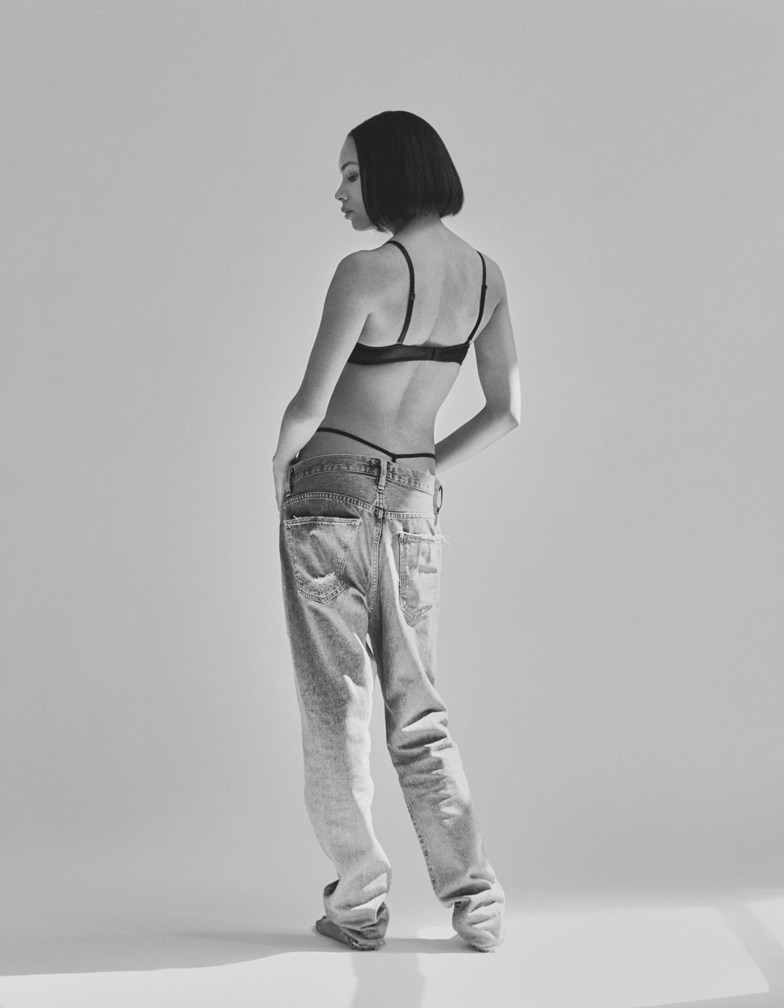 Symon seen from behind in a black bra and low-slung oversized jeans, black and white