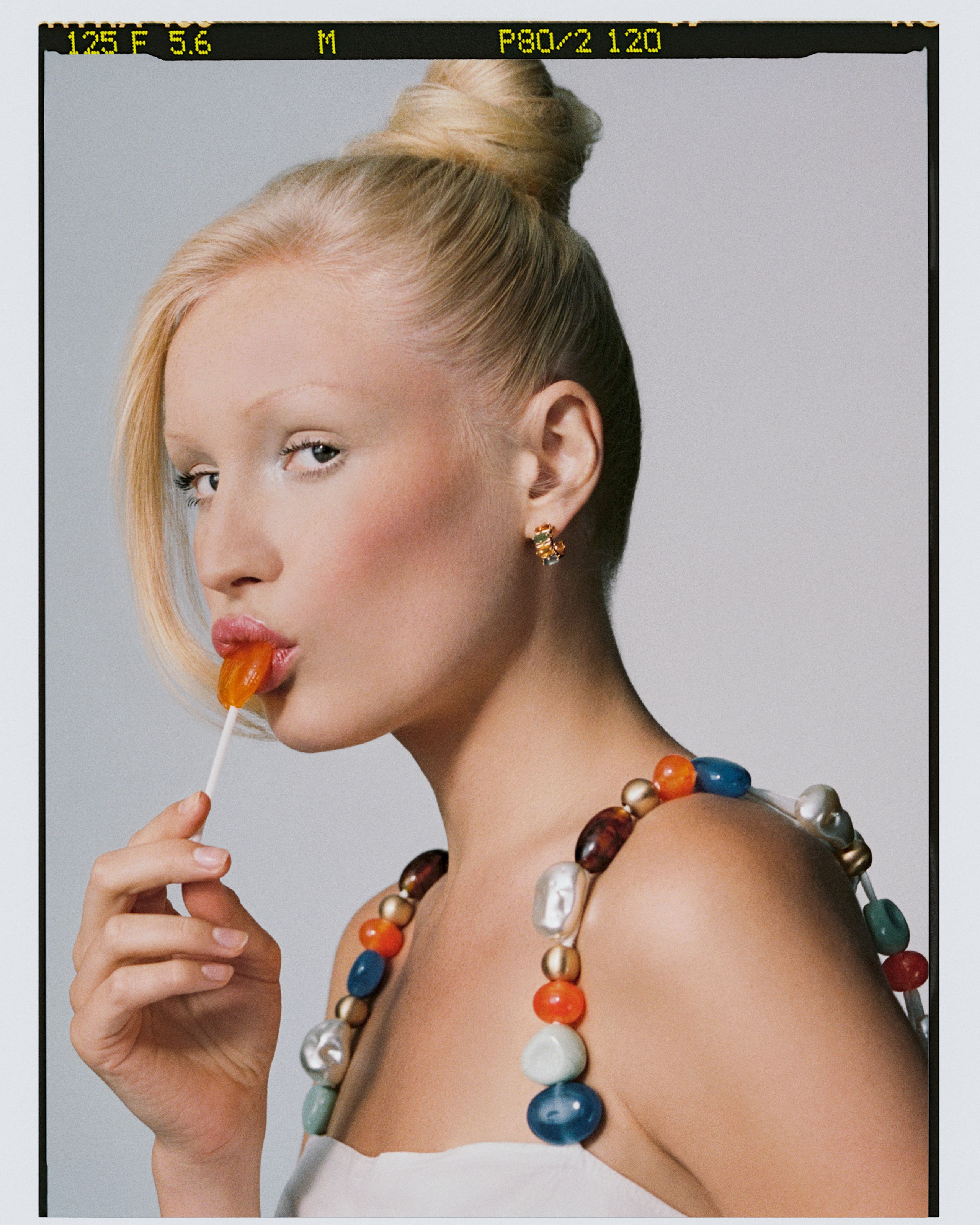 Film frame of a model with a top knot and an orange lollipop, oversized bead straps on bare shoulders