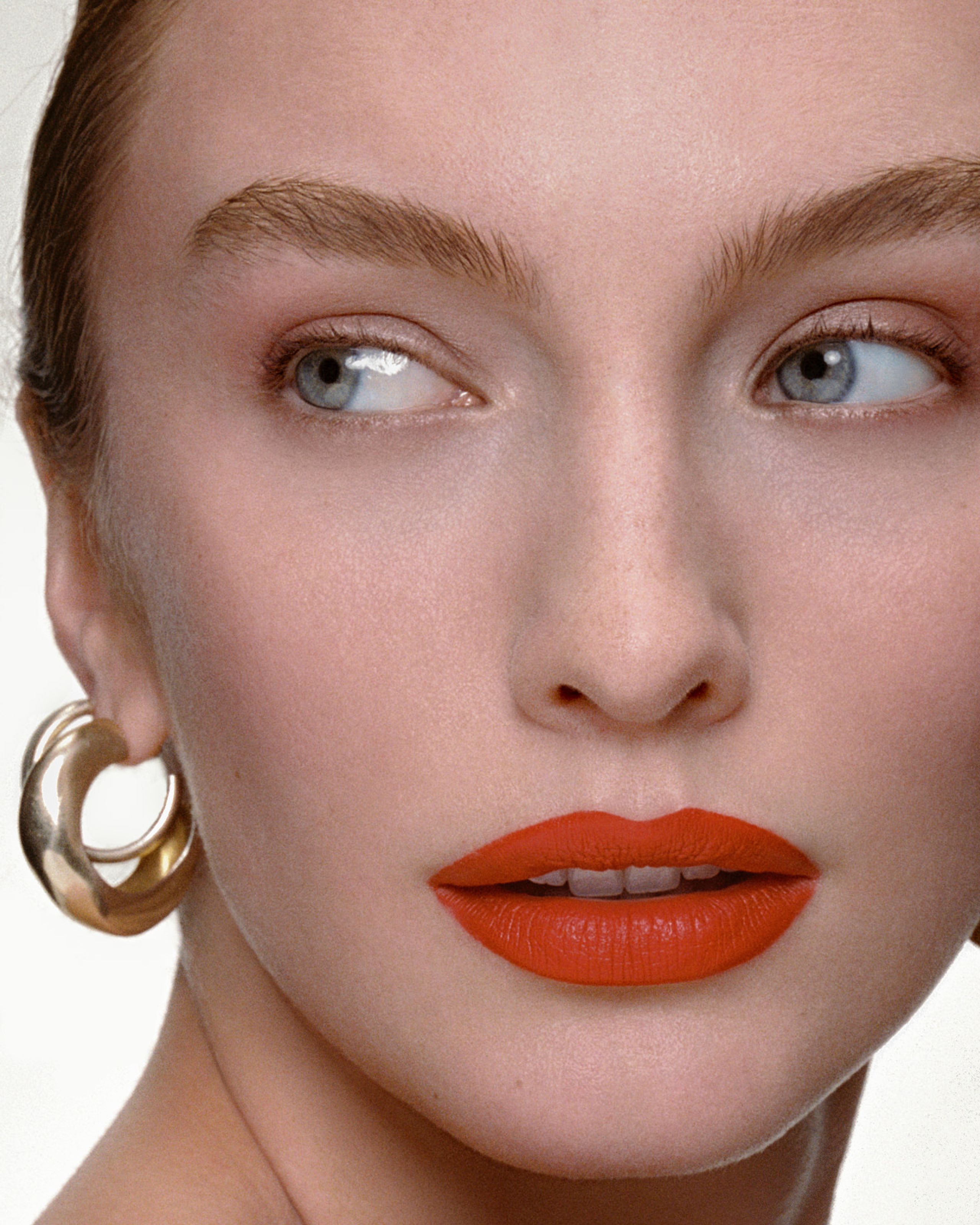 Direct gaze, feathered brows and glossy orange lips framed by gold AGMES hoop earrings