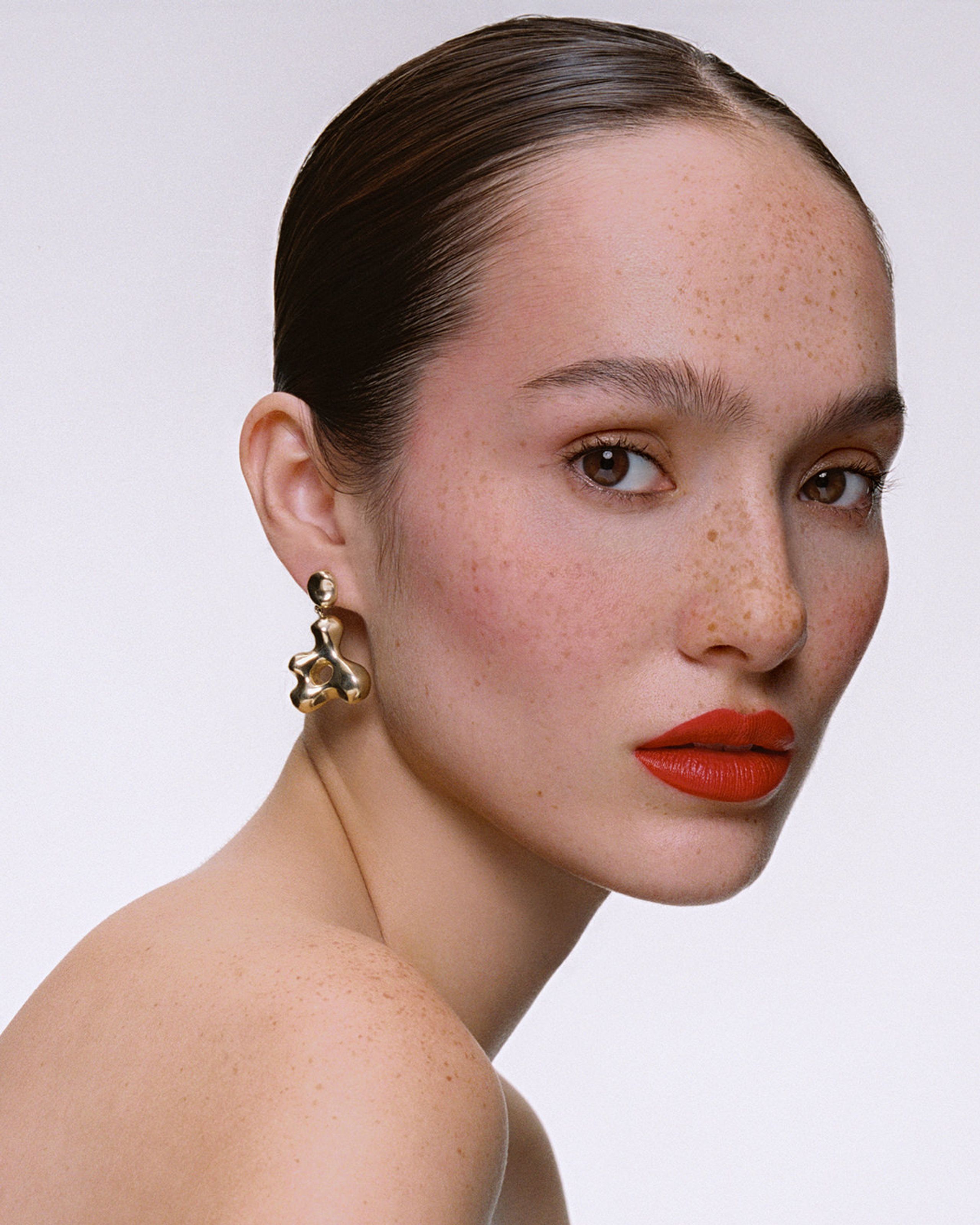 Siobhan with slicked-back hair glancing over a bare shoulder, red-orange lips and a gold AGMES earring