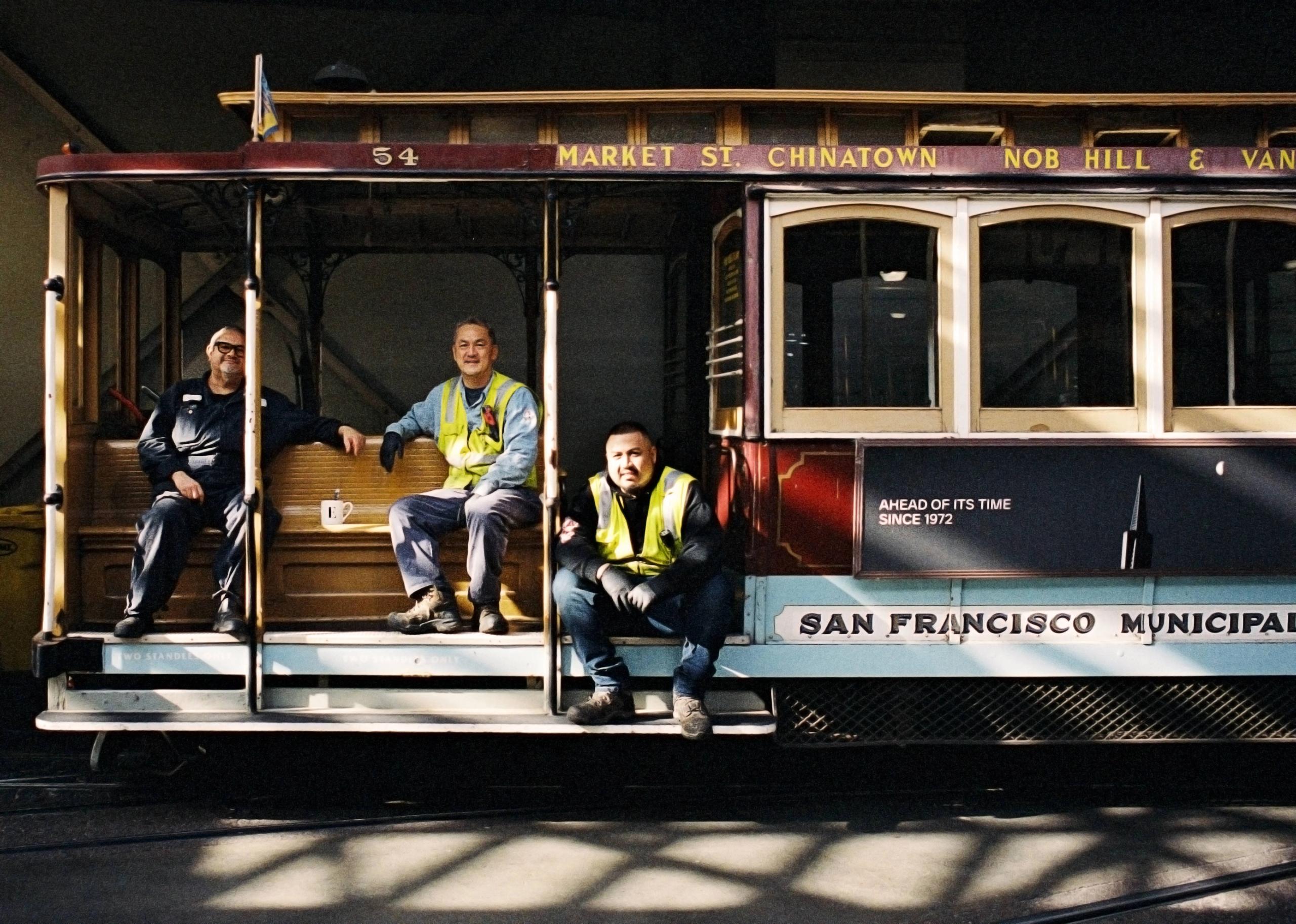 Three cable car workers in safety vests sitting on the running board of a cable car