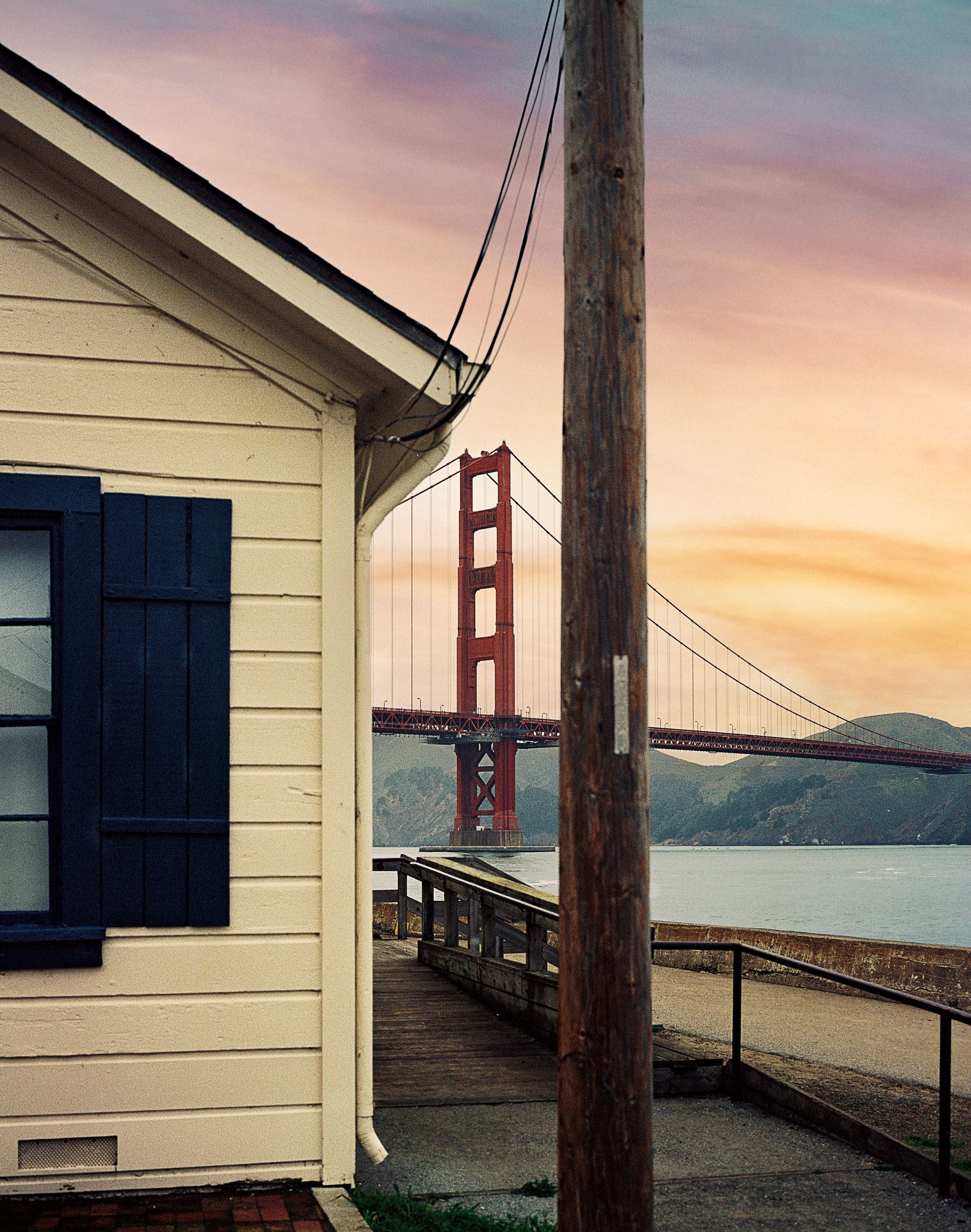 The corner of a clapboard building with blue shutters, the Golden Gate Bridge under a pink sky