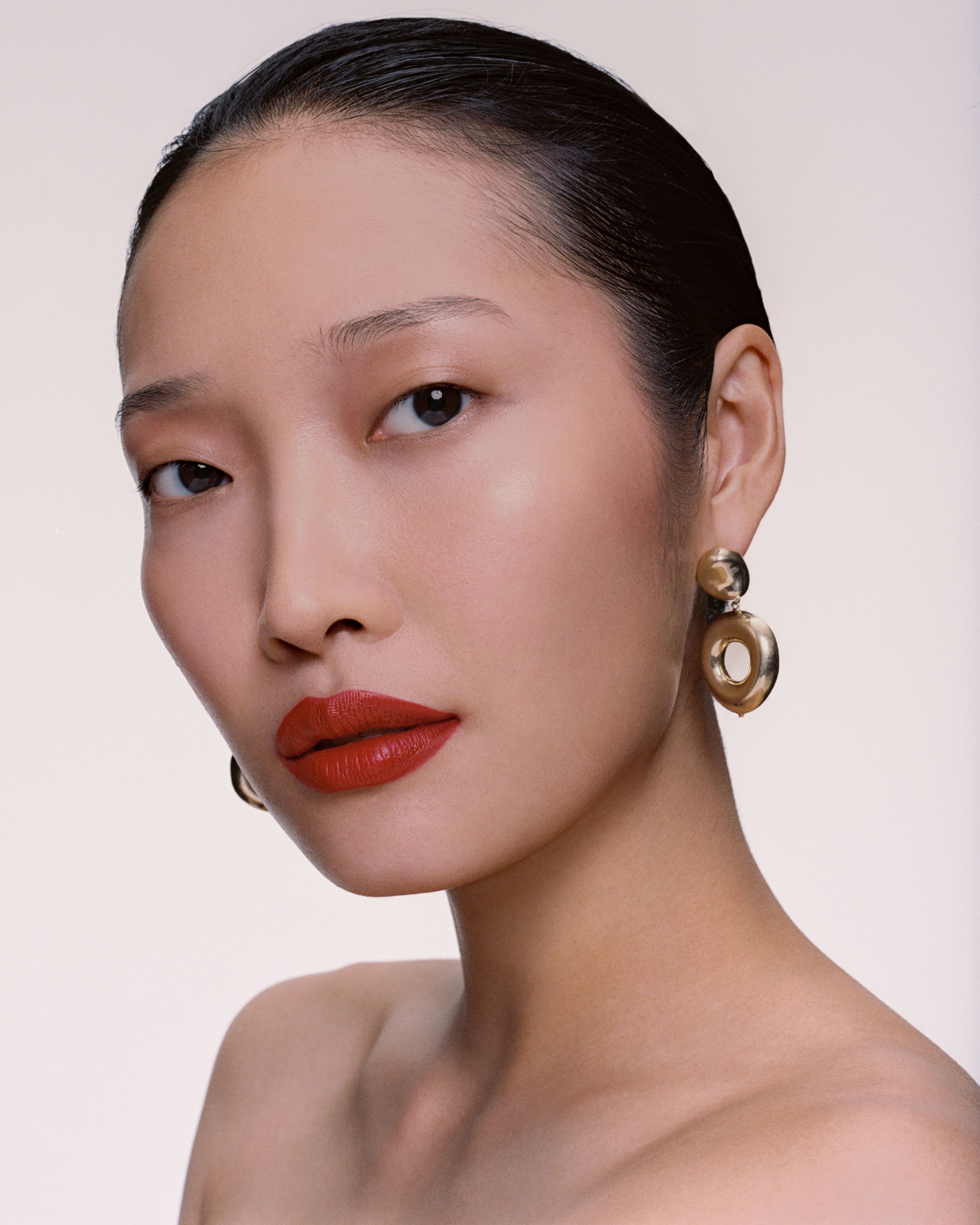 Model with slicked-back black hair and red-orange lips, gold AGMES earrings, turning toward the camera